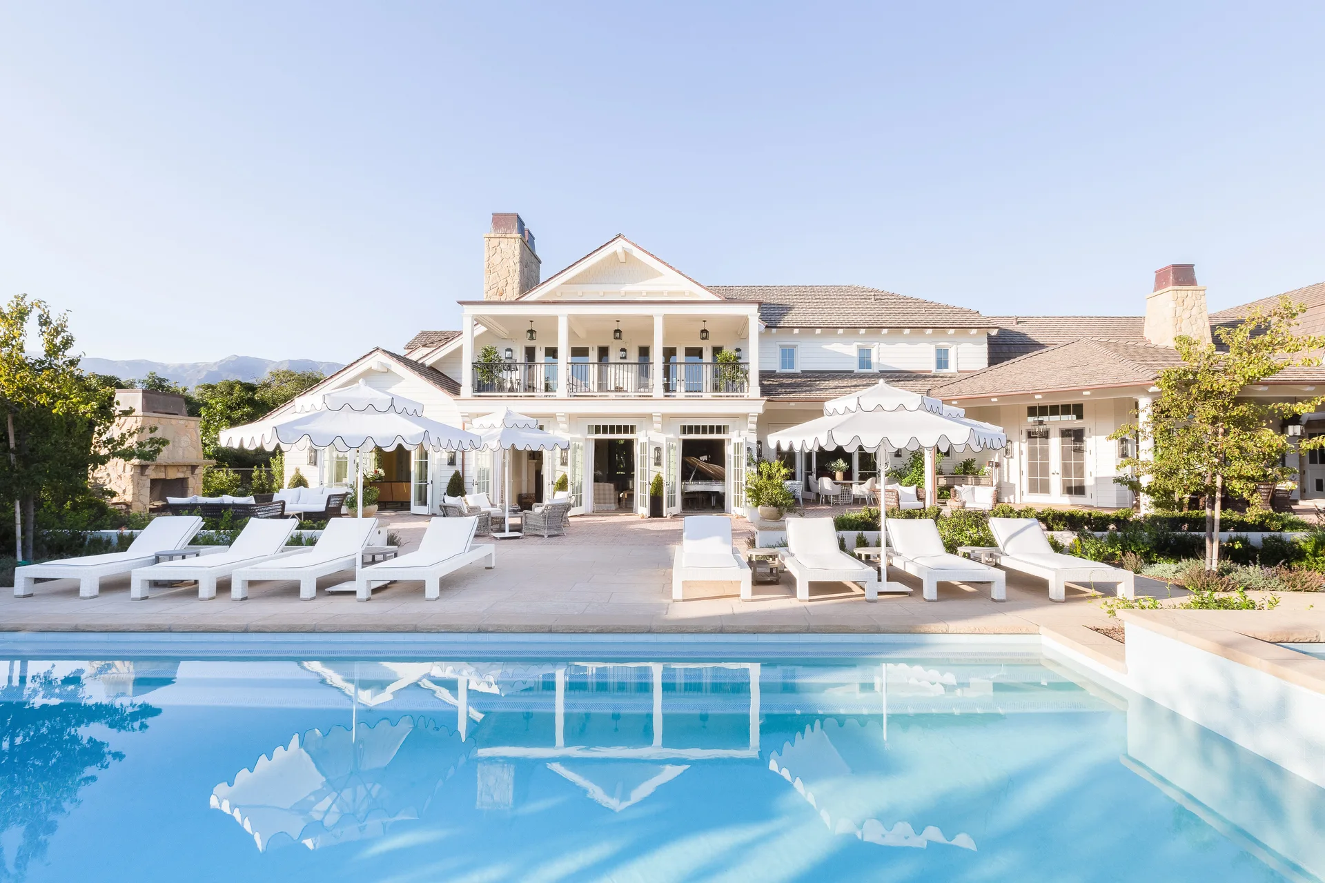 A swimming pool reflects white scalloped umbrellas and lounge chairs arranged on a stone patio in front of a white two-story house.