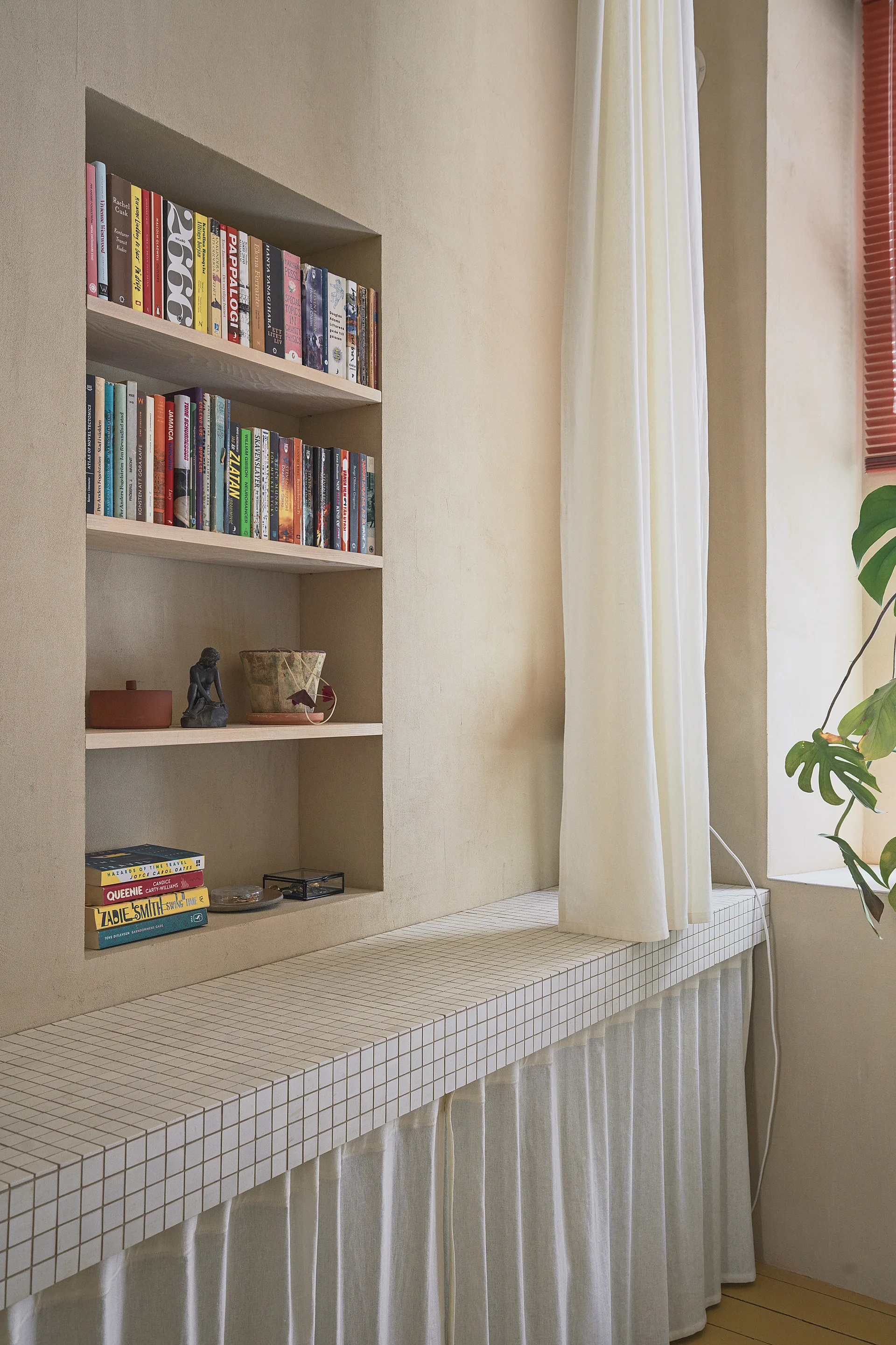 A recessed plaster wall niche with bookshelves sits above a counter covered in small square tiles with a gathered white fabric curtain skirt below.