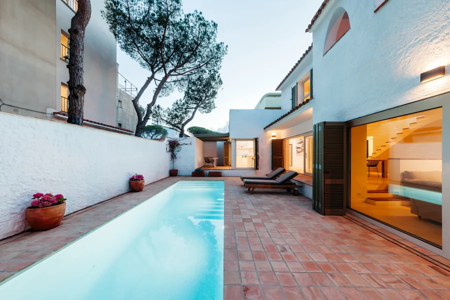 An elongated rectangular swimming pool is built next to a terracotta tile patio and a white stucco building with folding shutters.