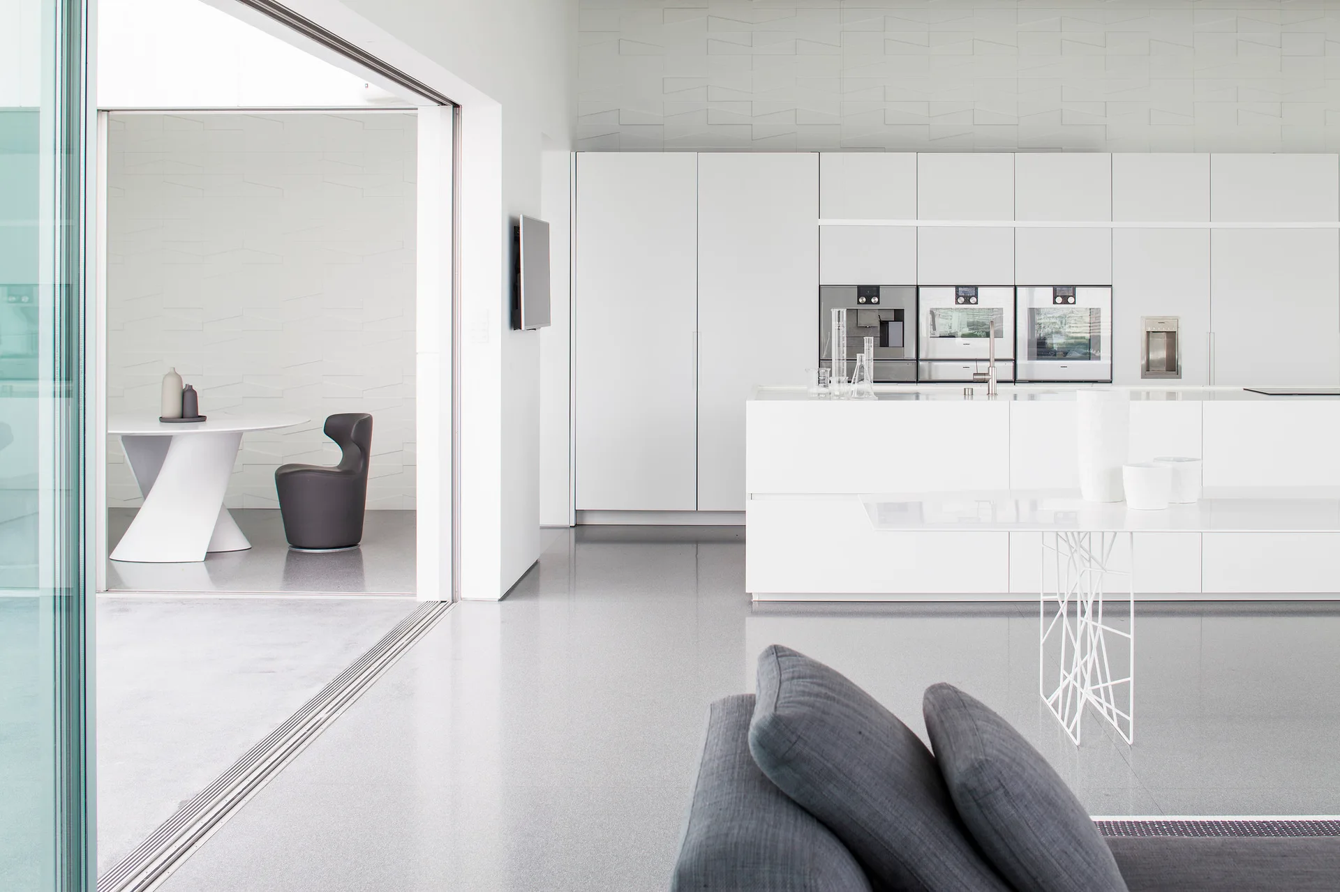 A minimalist, open-plan white kitchen features seamless integrated cabinetry, a central island, polished grey flooring, and a foreground grey sofa.