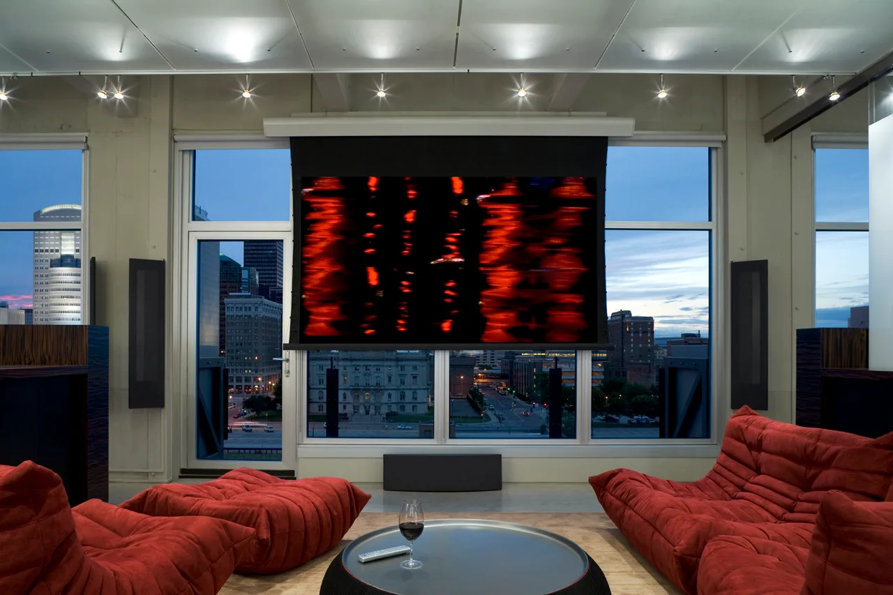 A modern media room features low-profile red tufted sofas, a dark coffee table, and a large projection screen in front of tall windows.