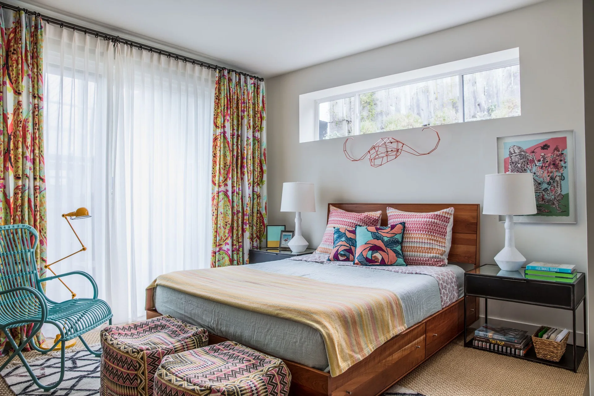 An eclectic bedroom features a wooden platform bed with colorful pillows, a high transom window, and a teal rocking chair.