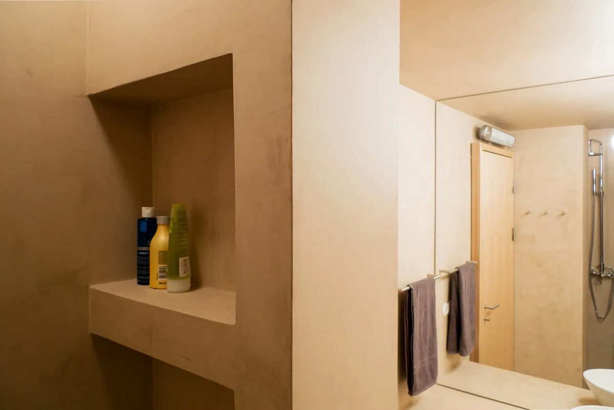 A minimalist bathroom featuring beige plaster walls with a built-in recessed storage shelf, a large mirror, and brown towels hanging on a metal rack.