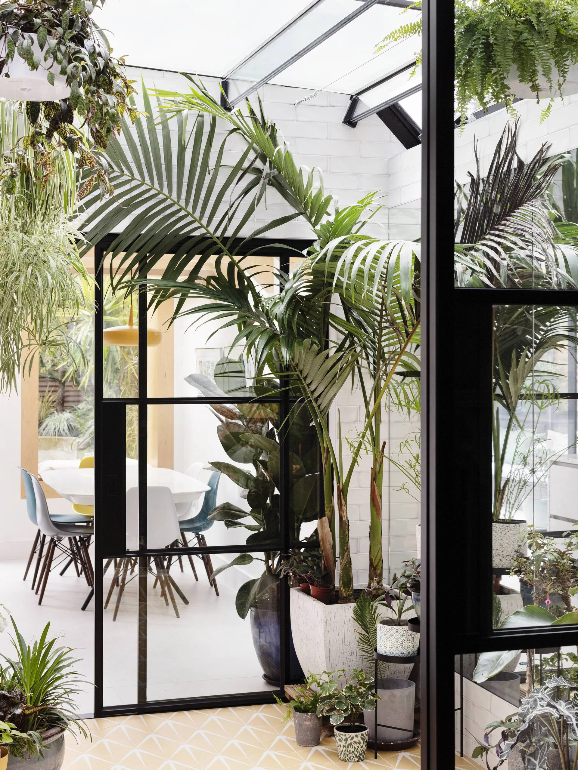 A bright conservatory features black-framed glass doors, a glass ceiling, lush indoor plants, and a dining table with chairs in the background.
