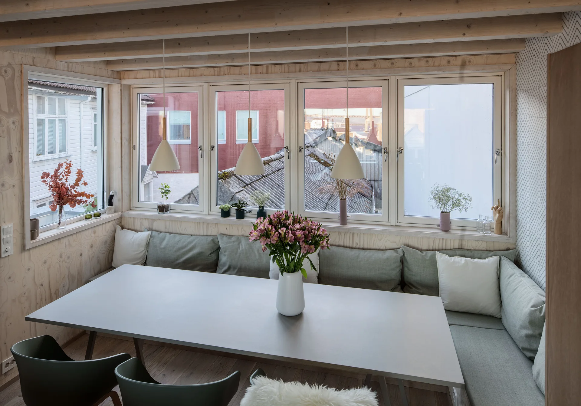 A bright dining nook features a grey table, an L-shaped banquette with green cushions, plywood walls, and three white pendant lights.