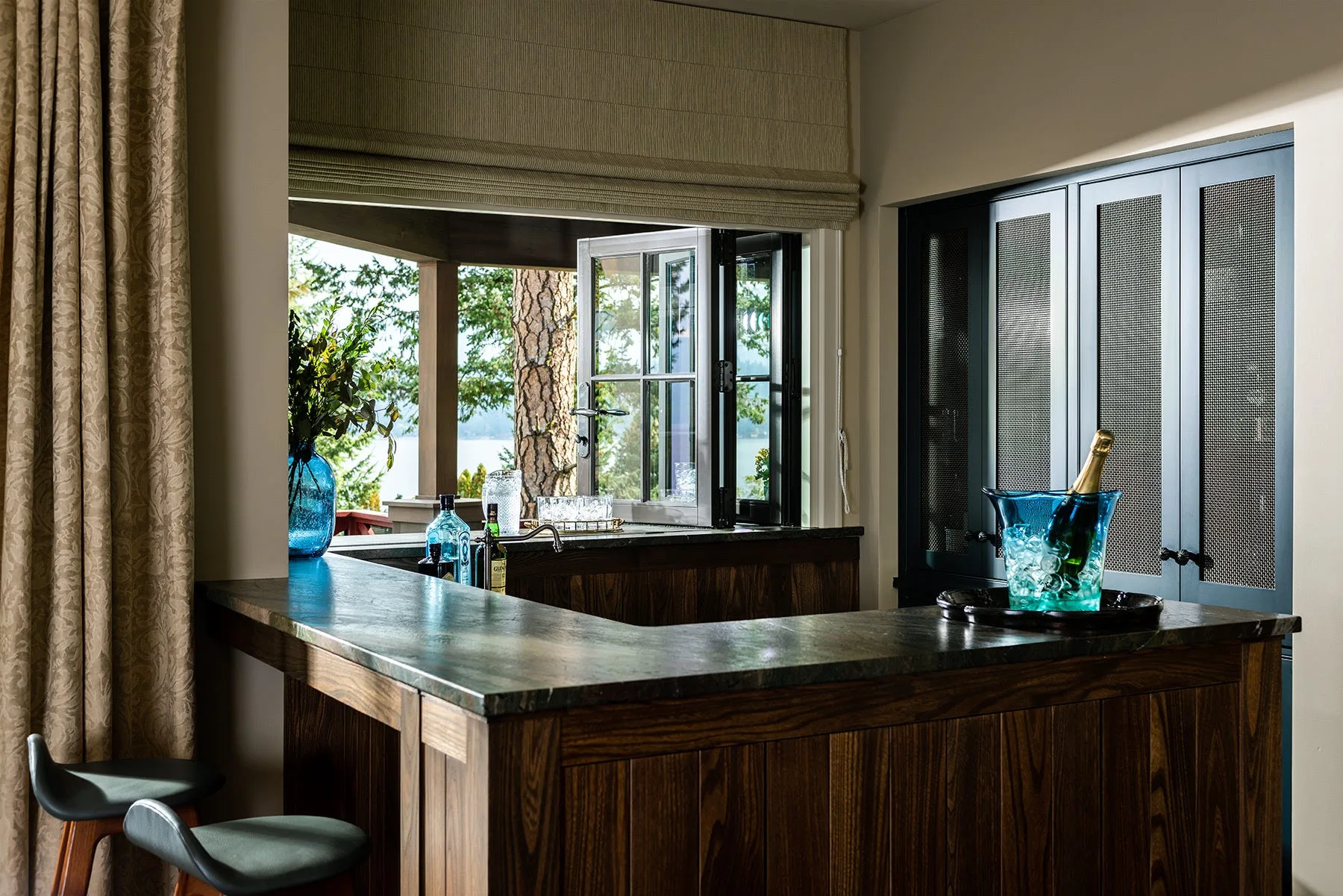 A dark wood home bar with a stone countertop, featuring a blue glass vase and ice bucket near a window overlooking a lake.