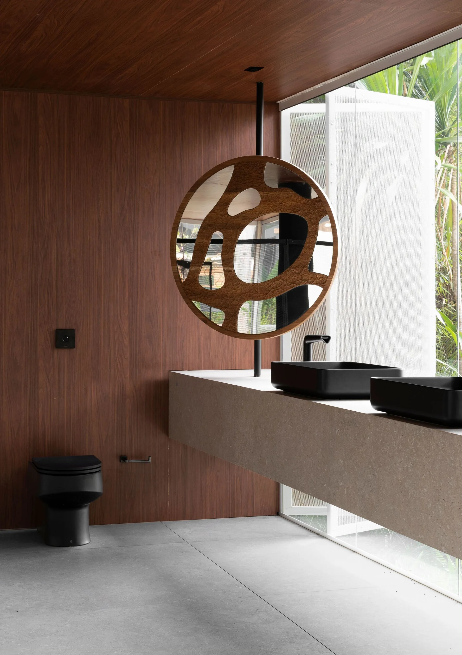 A minimalist bathroom with dark wood-paneled walls, a floating concrete vanity with black sinks, a circular mirror, and a black toilet.