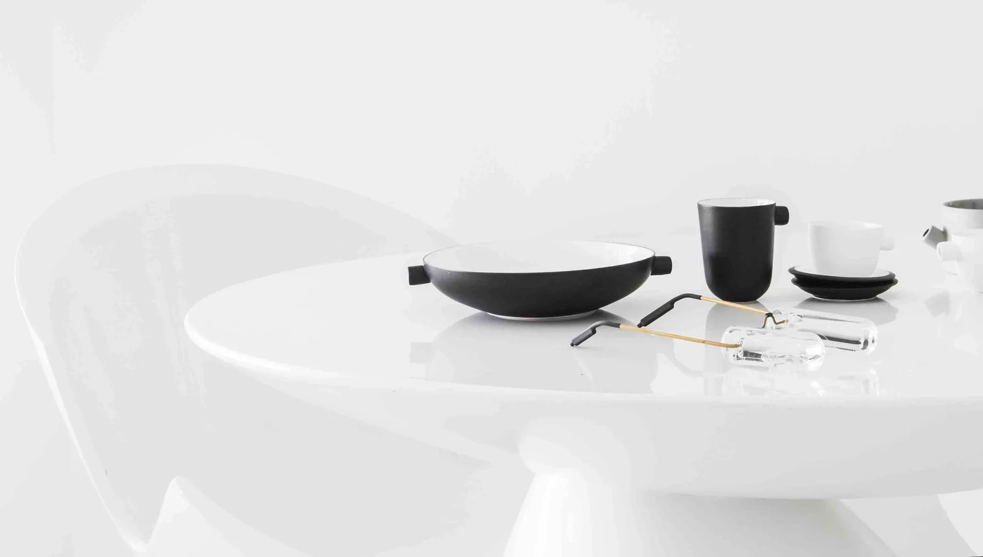 A glossy white round dining table features minimalist black and white ceramic tableware next to a pair of clear eyeglasses against a white background.