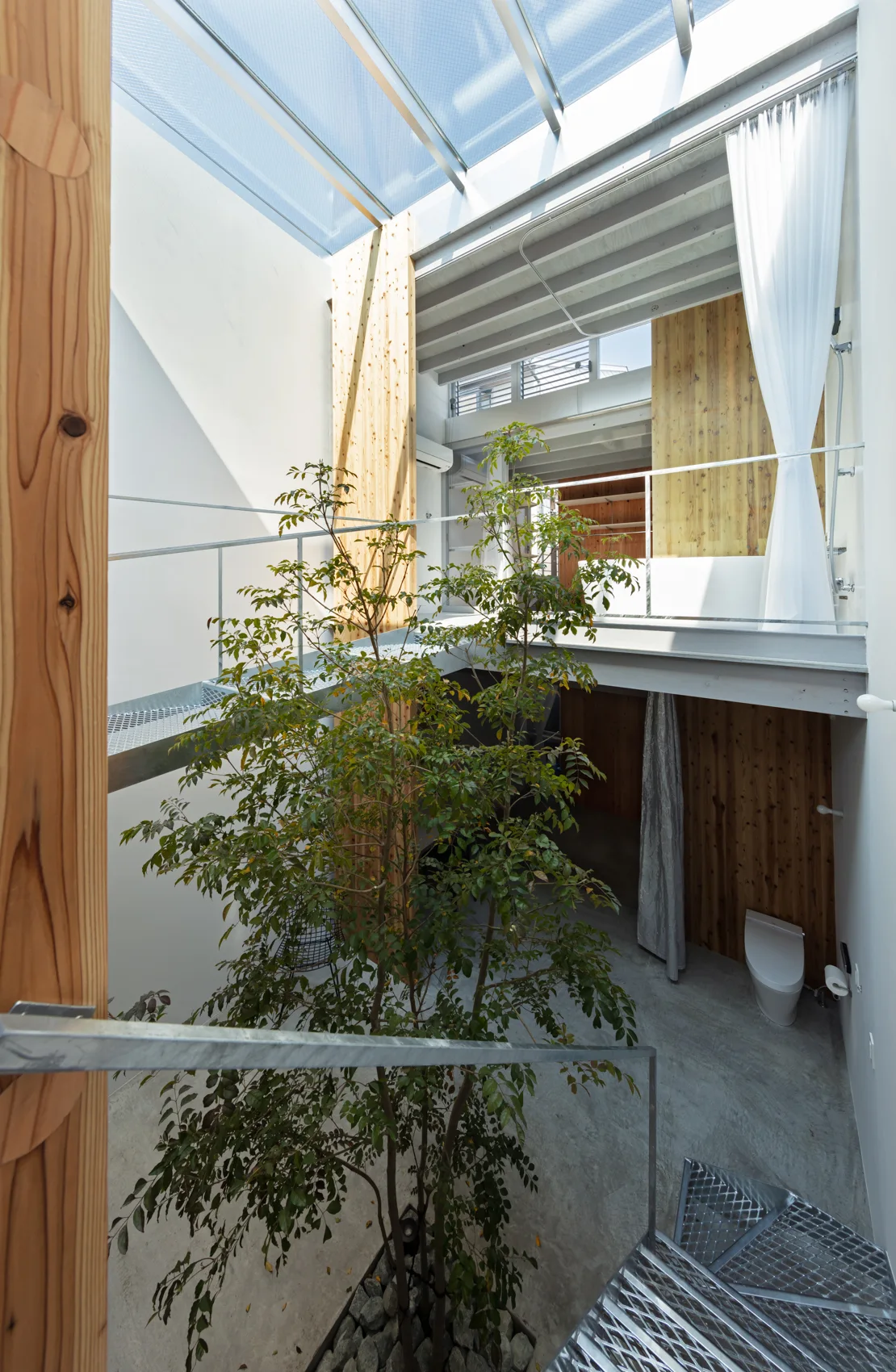 A multi-level minimalist atrium features a tall leafy indoor tree, metal grate walkways, natural wood paneling, and a large overhead glass skylight.