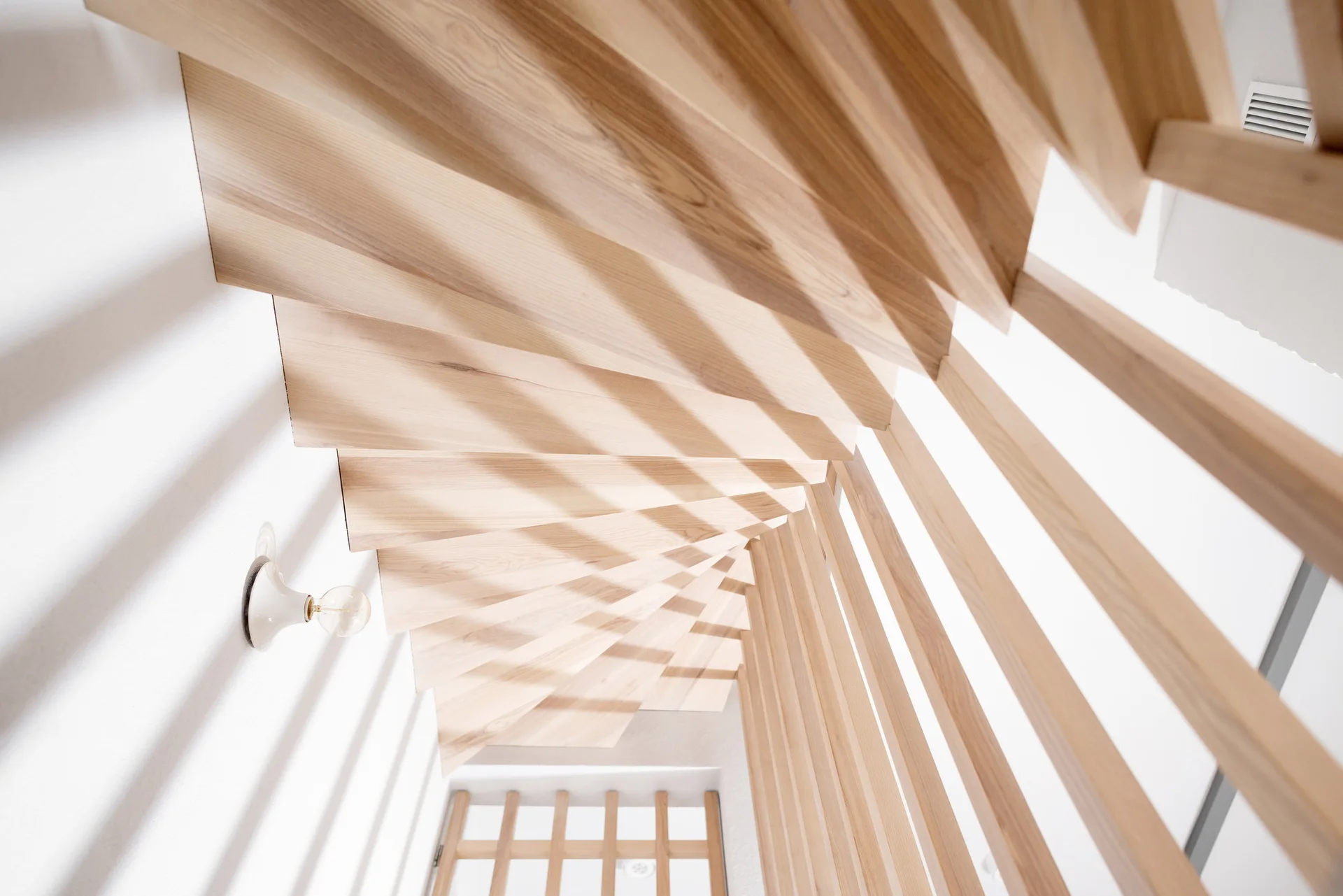 A high-angle view of a light wood staircase with vertical slat railings and a white wall with a small light fixture.