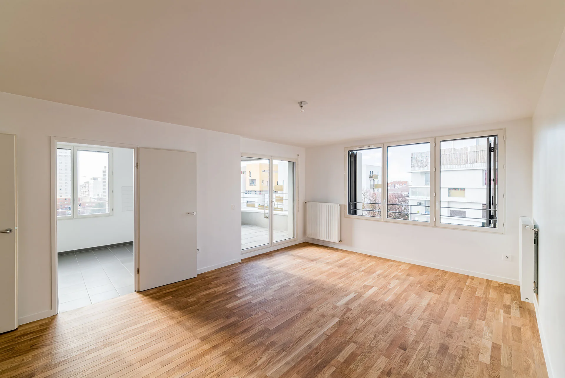 An empty living room features polished hardwood flooring, bright white walls, a balcony glass door, and large urban-facing windows.