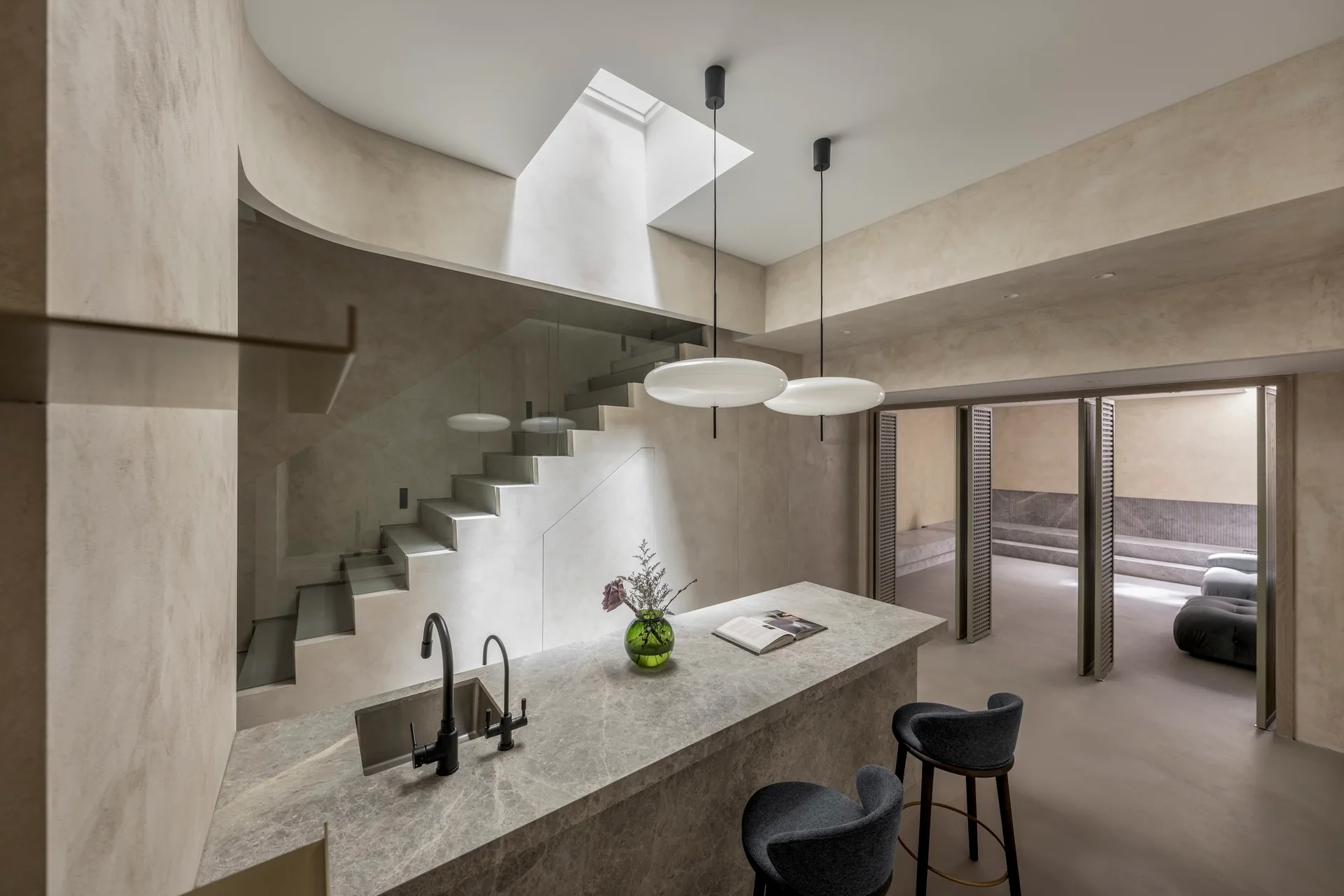 A minimalist kitchen features a beige stone island with black faucets, dark bar stools, and a glass-enclosed staircase beneath an overhead skylight.