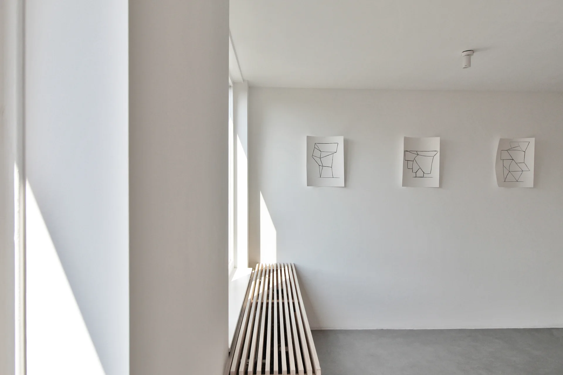 A minimalist hallway features a light wood slatted bench beneath a window, a grey concrete-like floor, and three unframed line drawings on a white wall.