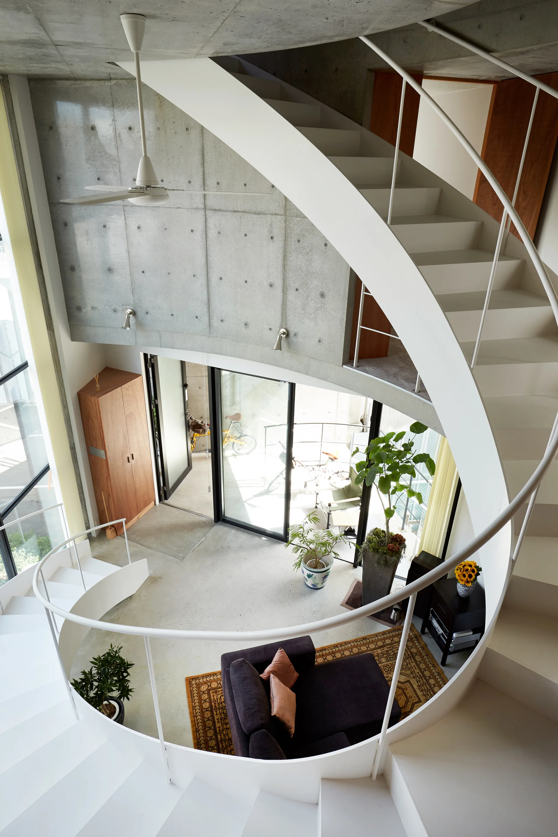 A high-angle view of a white spiral staircase curving over a minimalist living room with raw concrete walls and a dark sofa.