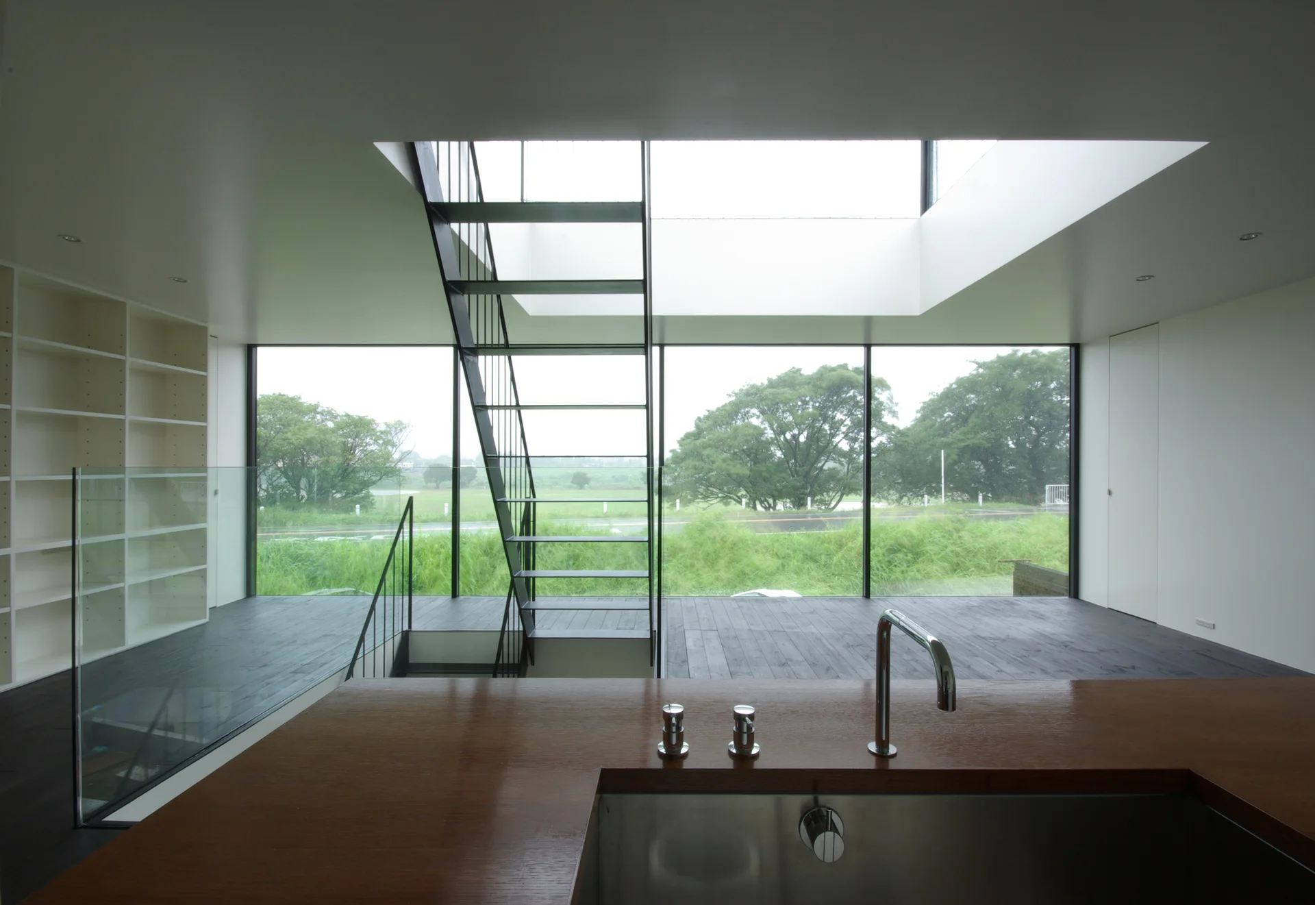 A modern kitchen foregrounds a wood island countertop, looking past an open metal staircase to a large glass wall displaying a green landscape.