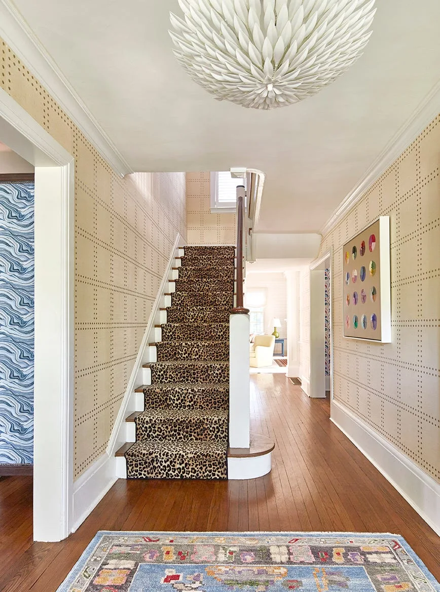 A bright foyer featuring a wooden staircase with a leopard-print runner, beige dotted wallpaper, and a white sculptural ceiling light.