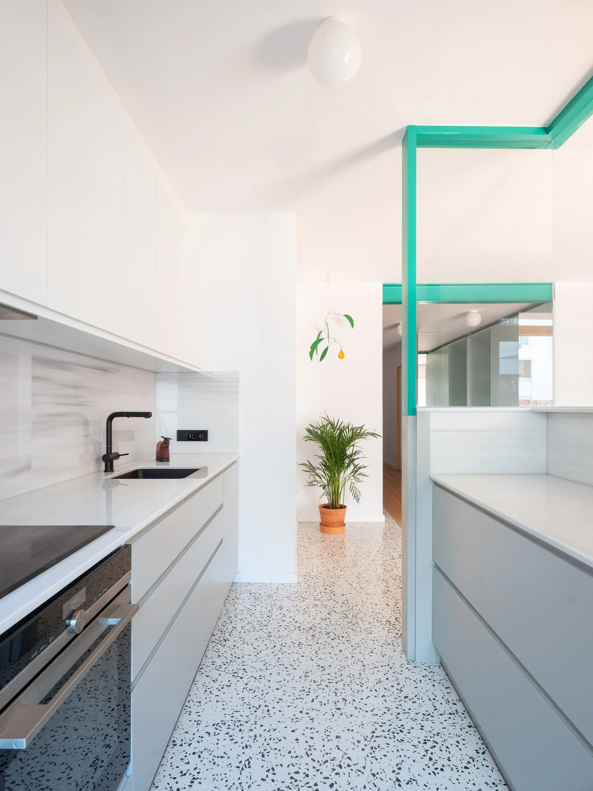 A contemporary kitchen featuring white and grey flat-front cabinets, speckled terrazzo flooring, and a glass partition with a bright teal frame.