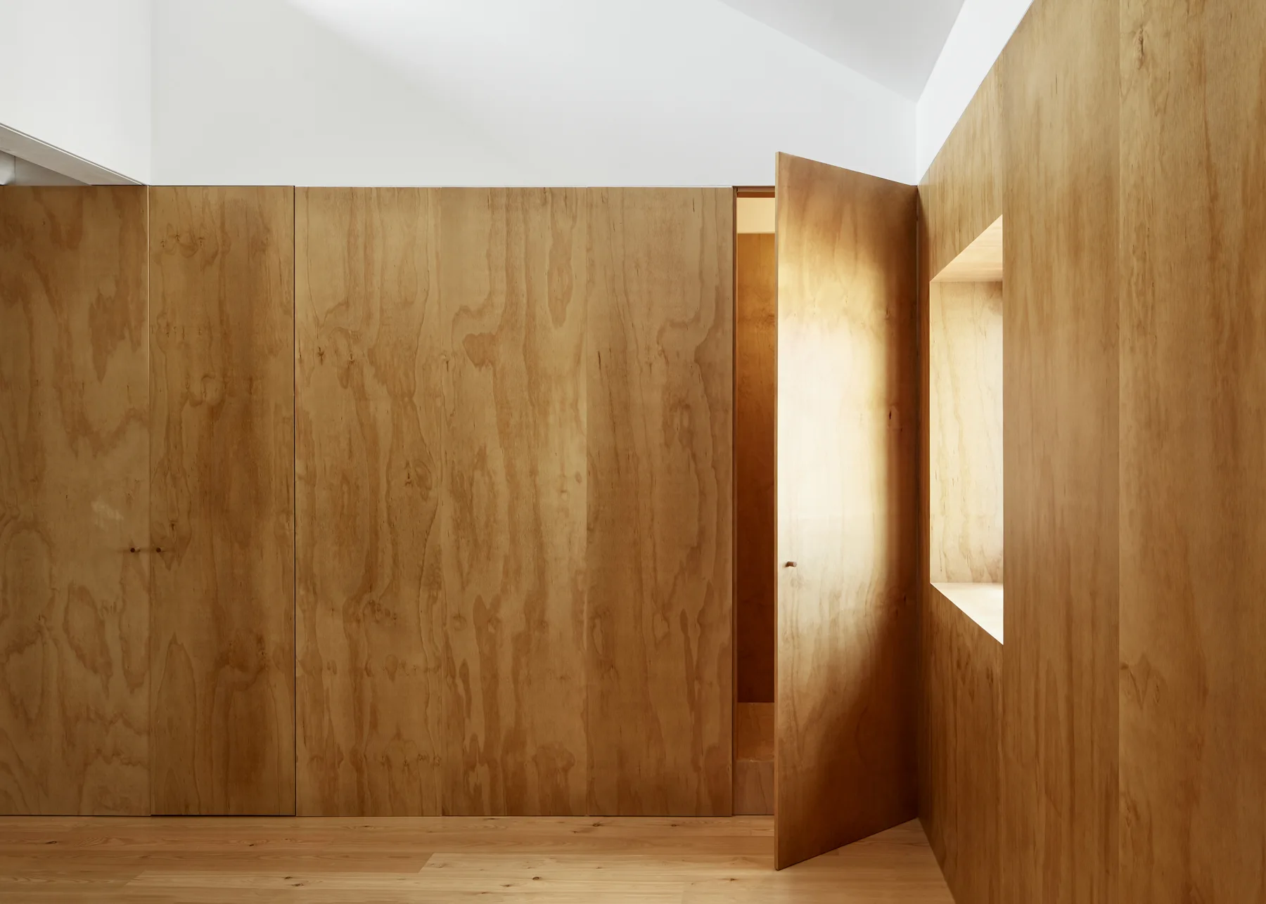 A modern minimalist interior with light wood floors and walls completely covered in plywood panels featuring a flush door and recessed window.