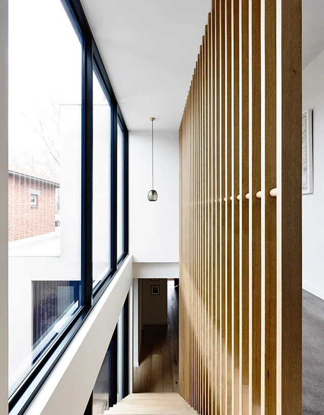 A modern staircase features a tall vertical wood slat screen next to large double-height windows.