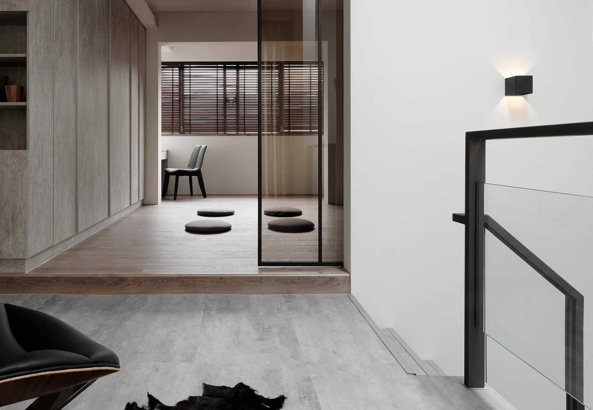 A modern minimalist staircase landing features a black glass railing, a wall sconce, and a raised wooden platform room with floor cushions.