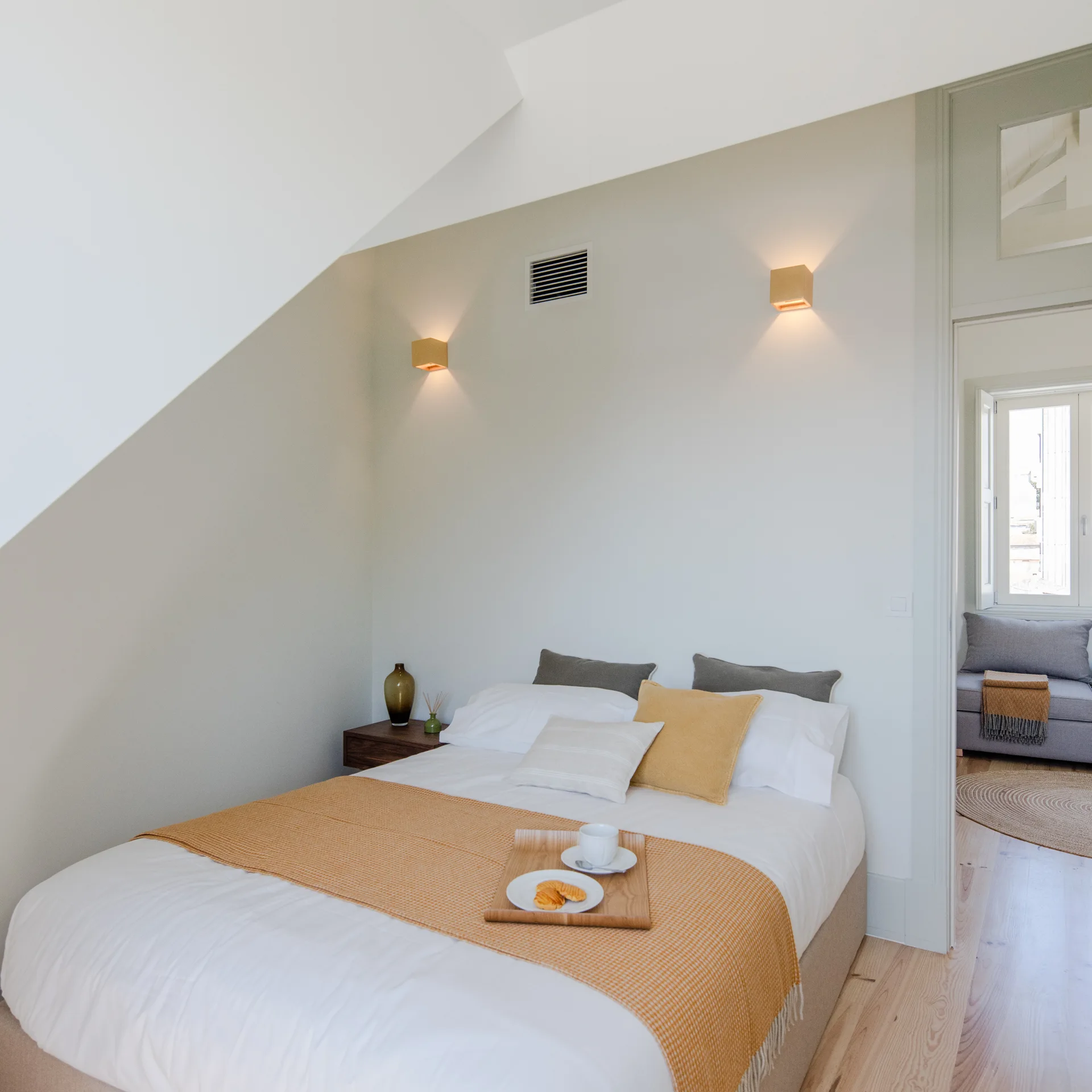 A minimalist attic bedroom with a low bed featuring mustard yellow and grey textiles, wooden wall sconces, and a sloped white ceiling.