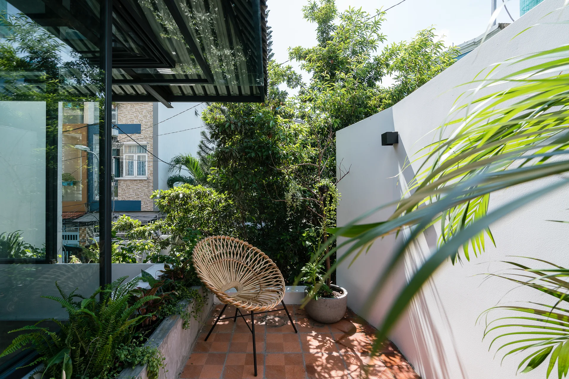 A woven circular rattan chair sits on a square terracotta tiled balcony next to lush green plants, a white wall, and a concrete planter.