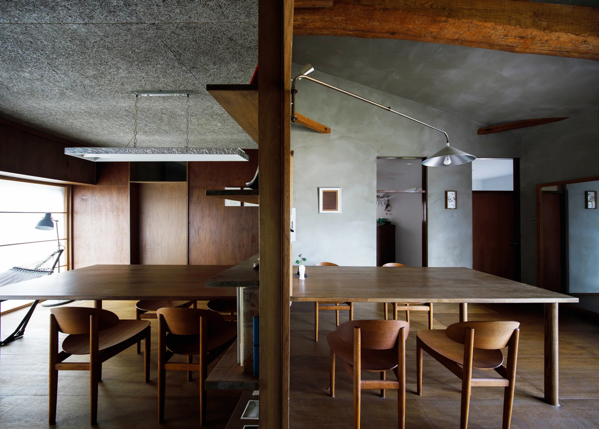 A minimalist dining room featuring two wooden tables separated by a central column partition under exposed beams and a textured ceiling.