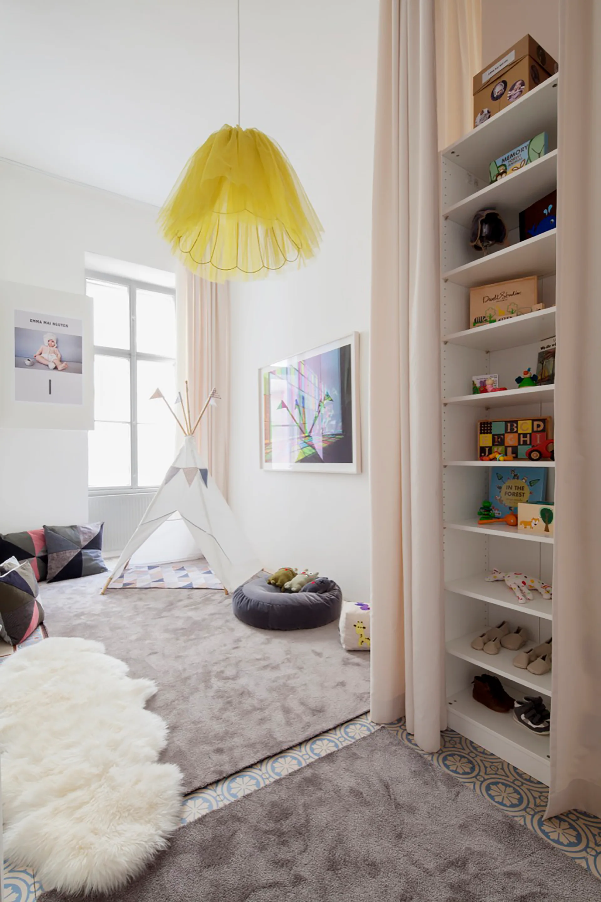 A light-filled kids playroom with white walls, a fabric teepee, a tall white bookcase framed by neutral drapes, and a yellow pendant light.