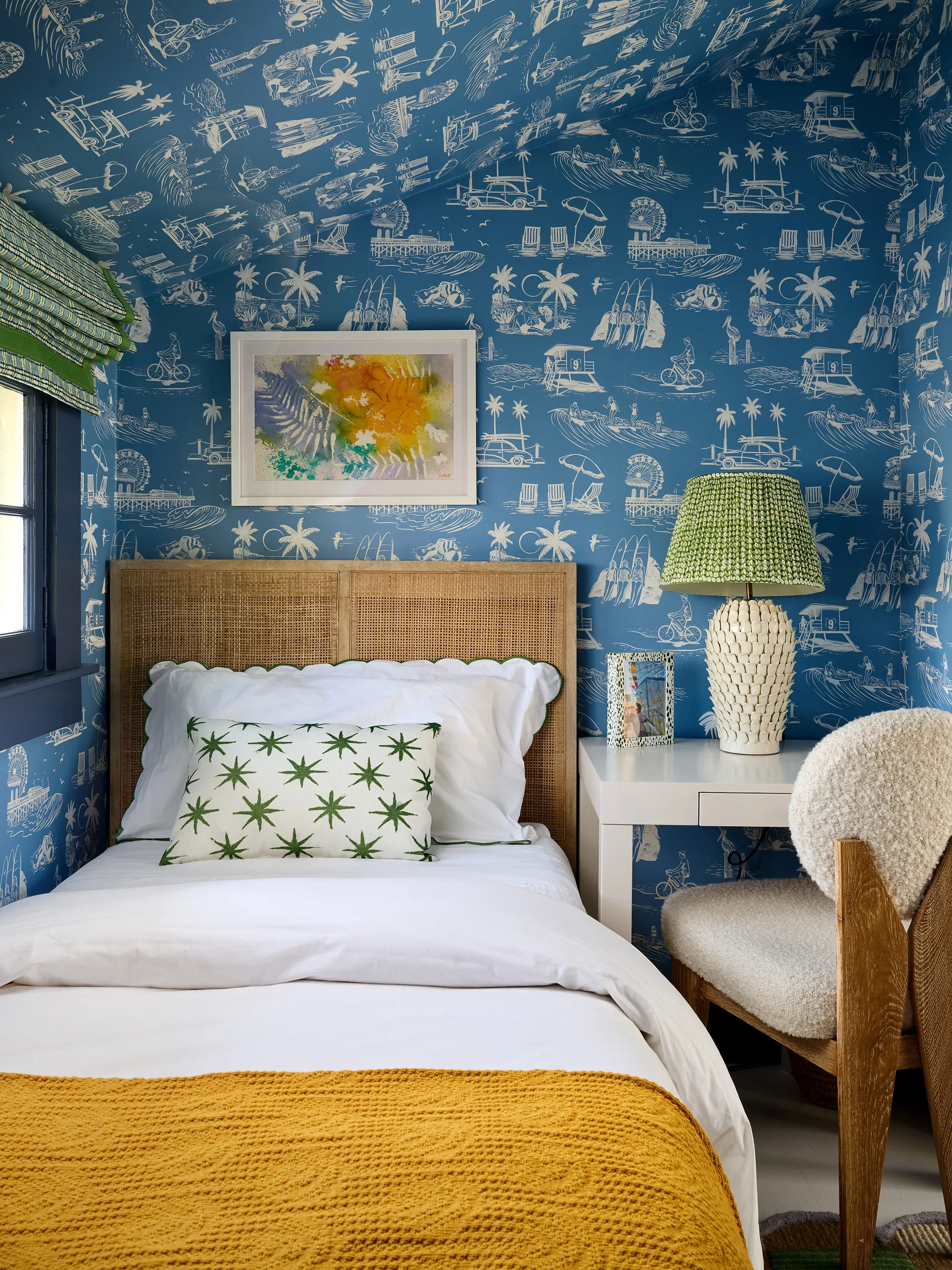 A cozy bedroom nook features blue coastal toile wallpaper on the walls and ceiling, a bed with a cane headboard, and a white writing desk.