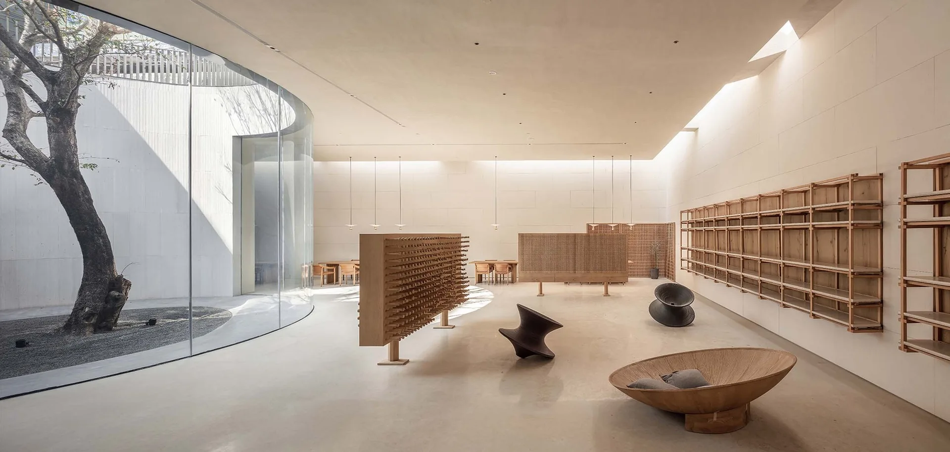 A minimalist interior space features a curved floor-to-ceiling glass wall overlooking a courtyard tree, wooden shelving, and sculptural lounge chairs.