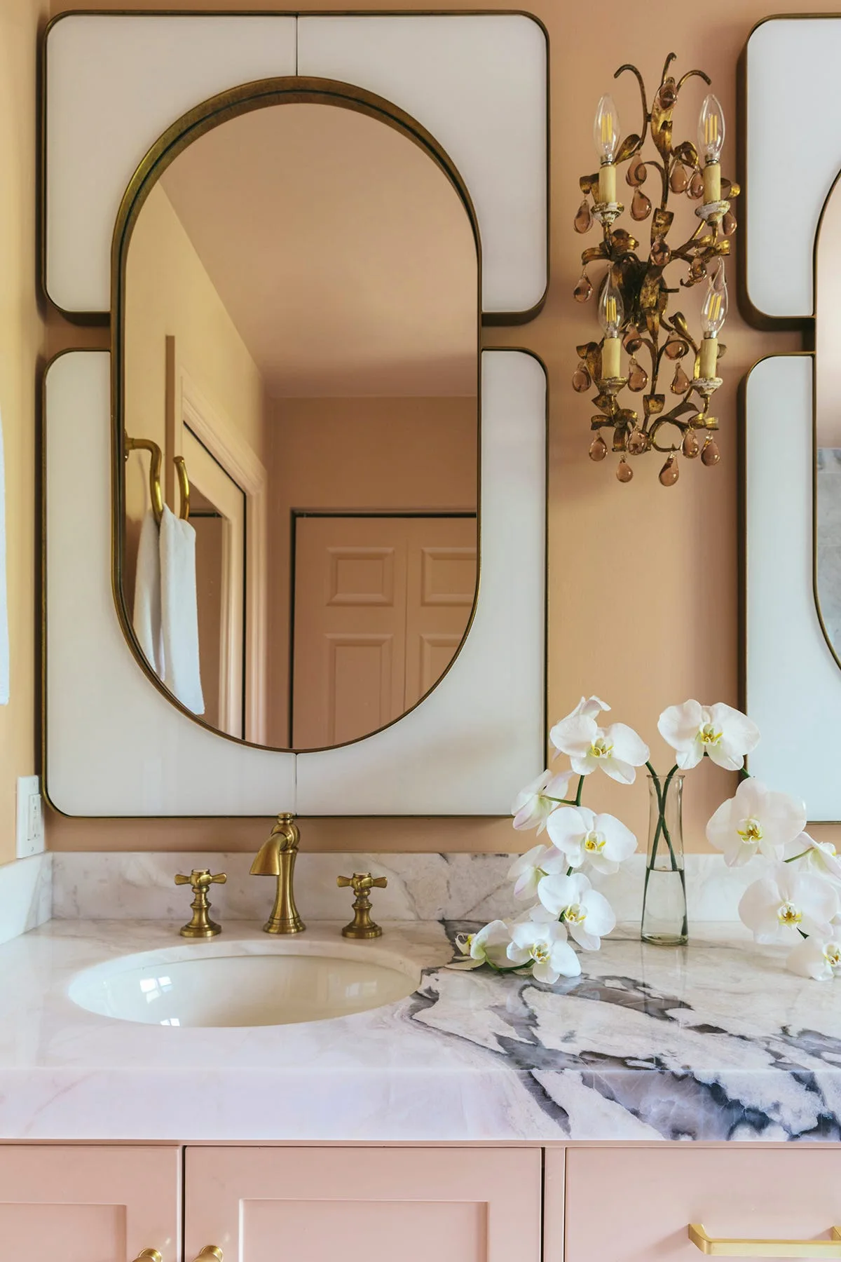 A bathroom vanity with a pink painted base, heavily veined white marble countertop, brass faucet, decorative mirror, and a brass wall sconce.