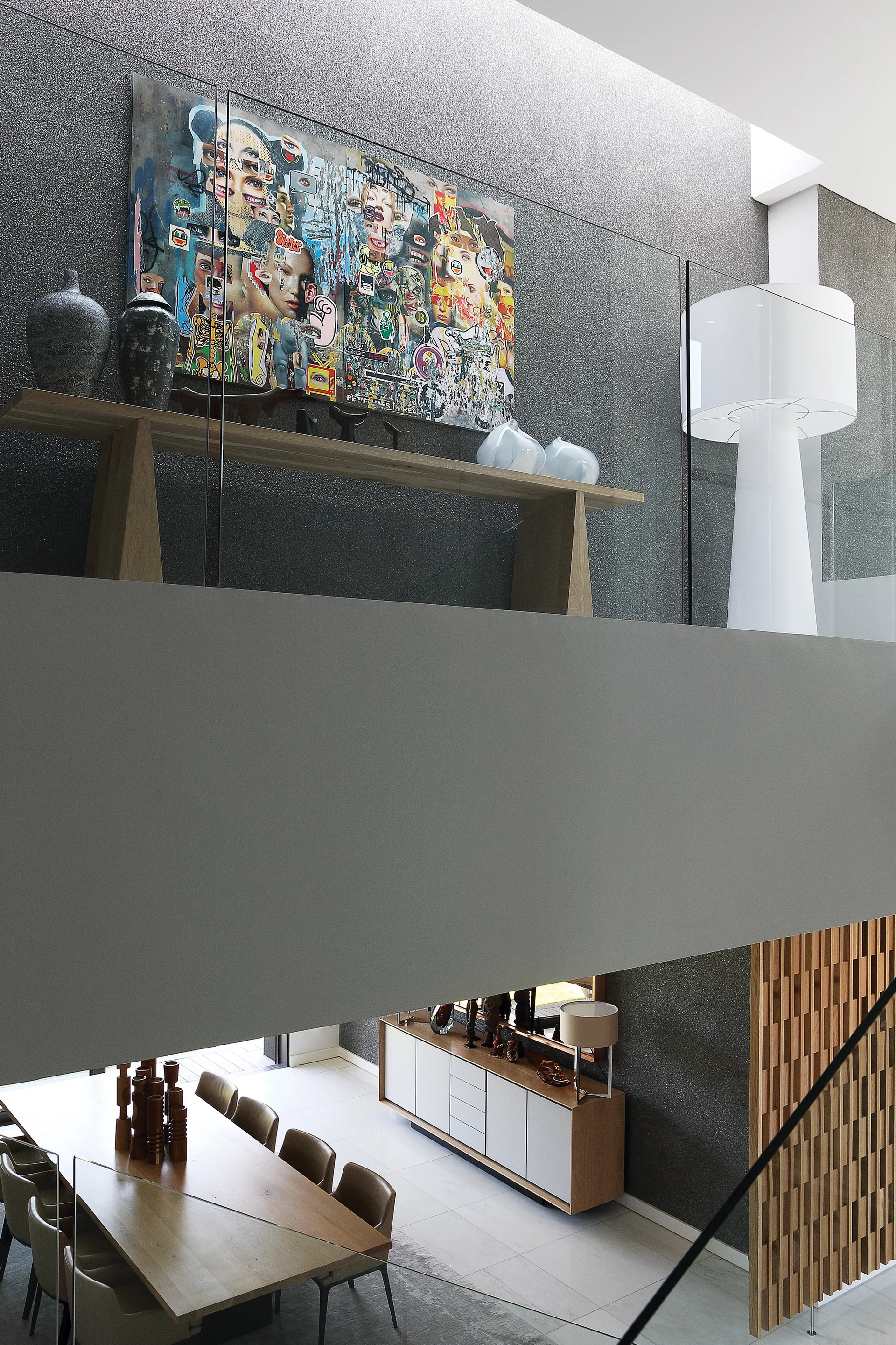 A modern split-level interior showing a dining room with a wooden table below and an upper landing featuring a glass railing, console table, and colorful art.