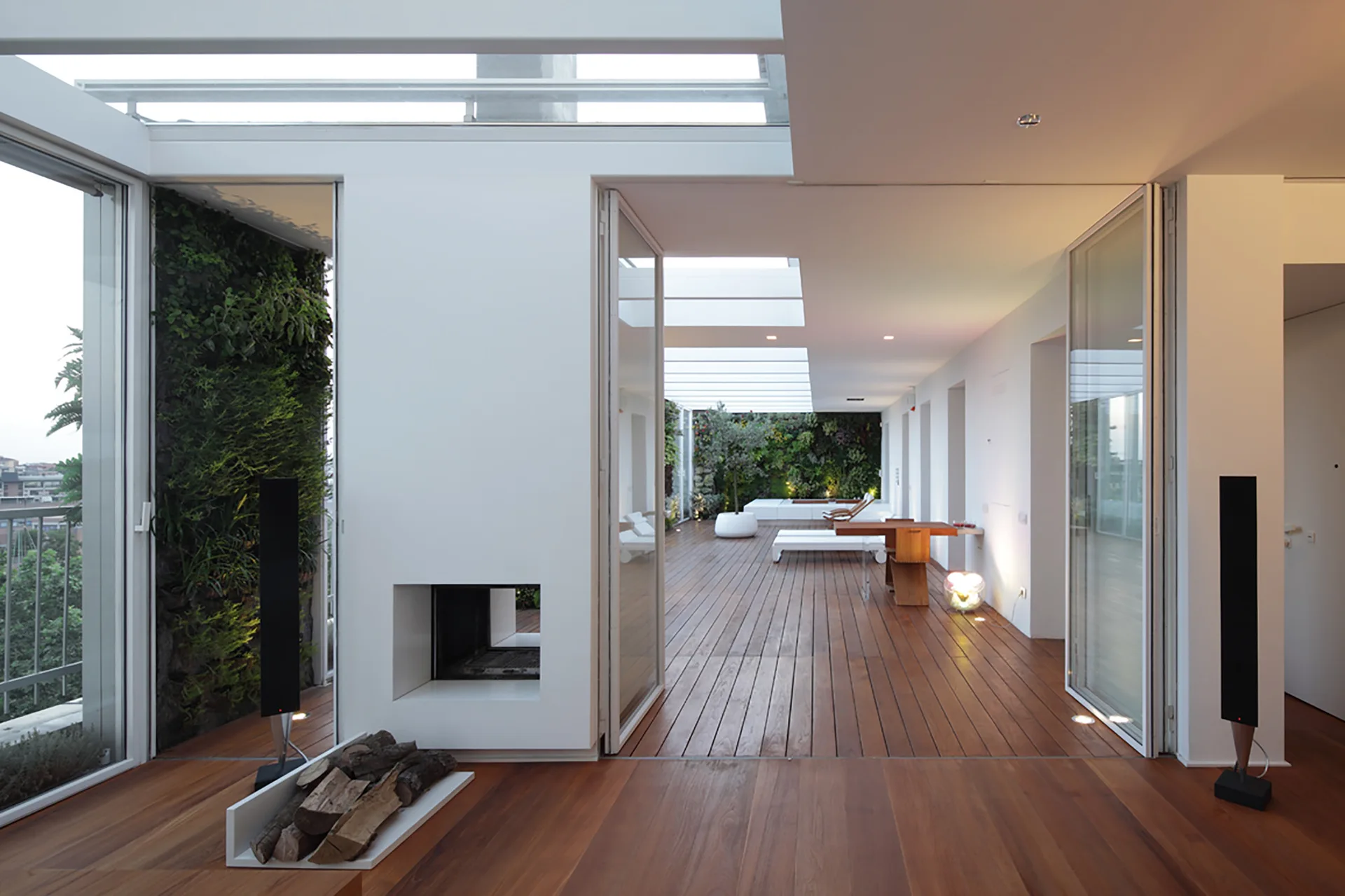 A modern open-plan indoor space connects to a large wooden deck terrace with a vertical living wall and retractable glass doors.