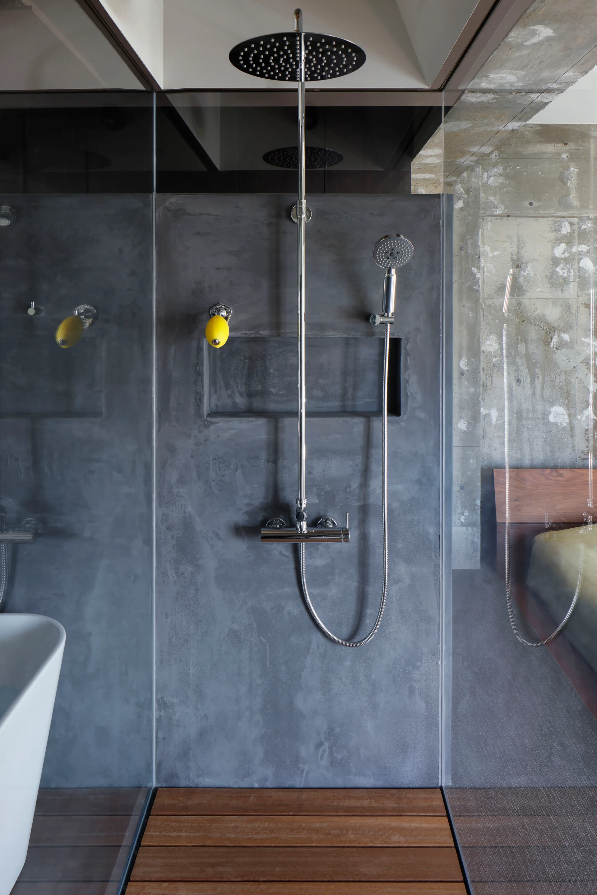 A minimalist shower featuring grey concrete walls, a chrome rain shower head, a recessed storage niche, and a slatted wood floor deck.