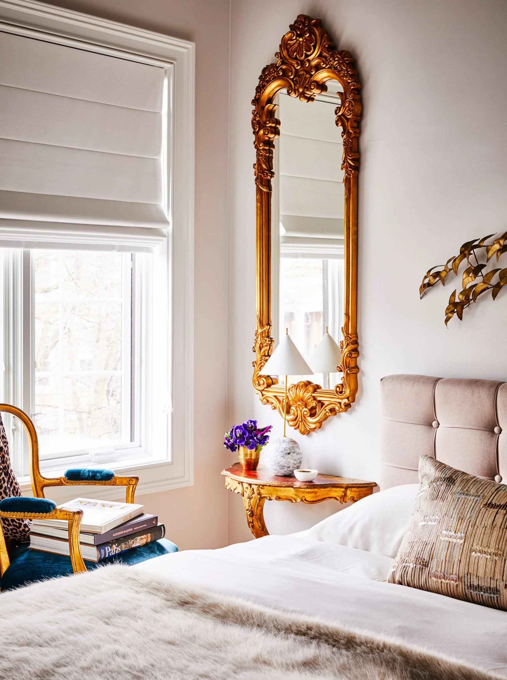 A bedroom corner features a bed with a tufted beige headboard, a tall ornate gold mirror, a gilded console table, and a blue velvet armchair by a window.