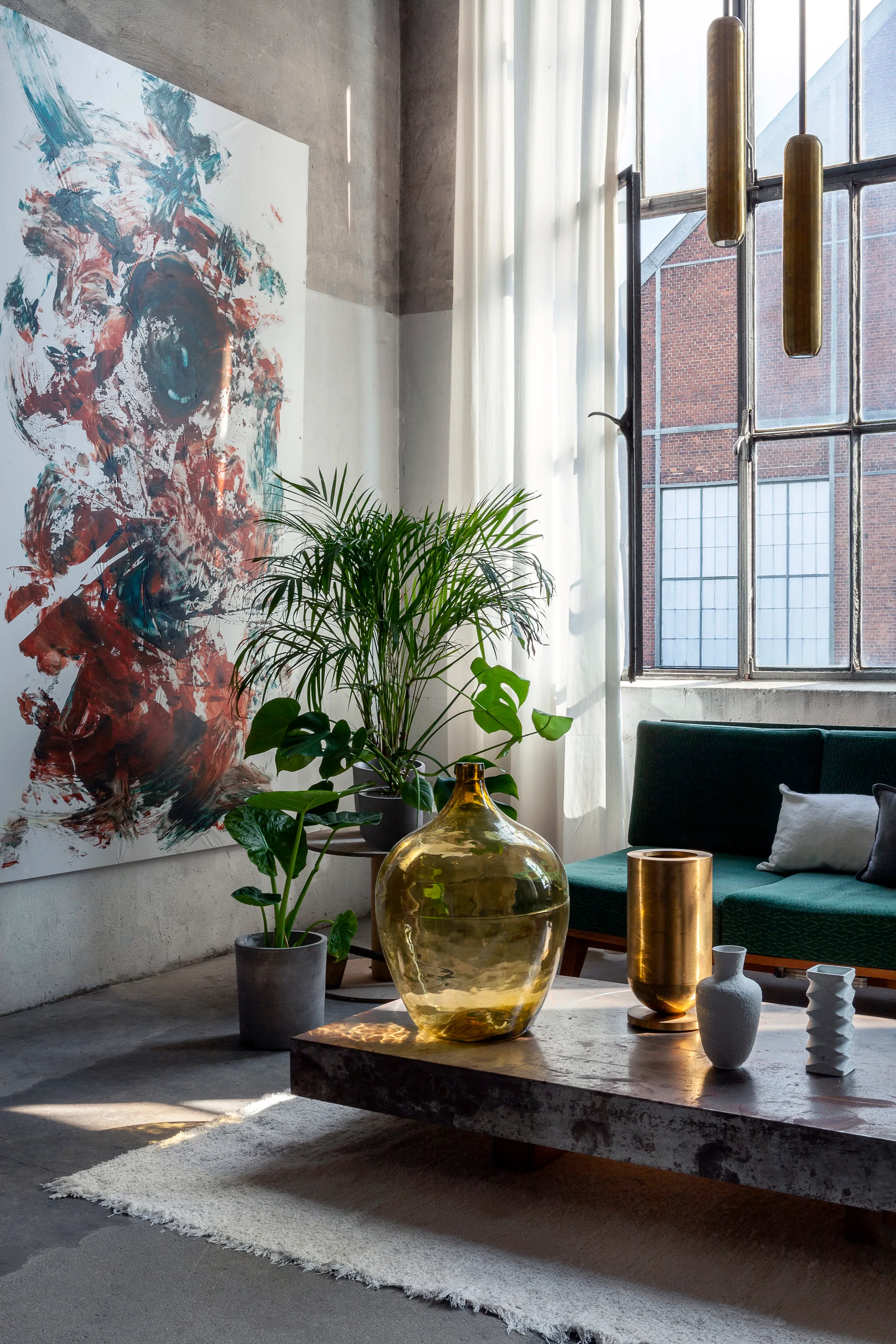 An industrial loft living room with a green sofa, weathered metal coffee table, potted plants, and a large abstract painting on a concrete wall.