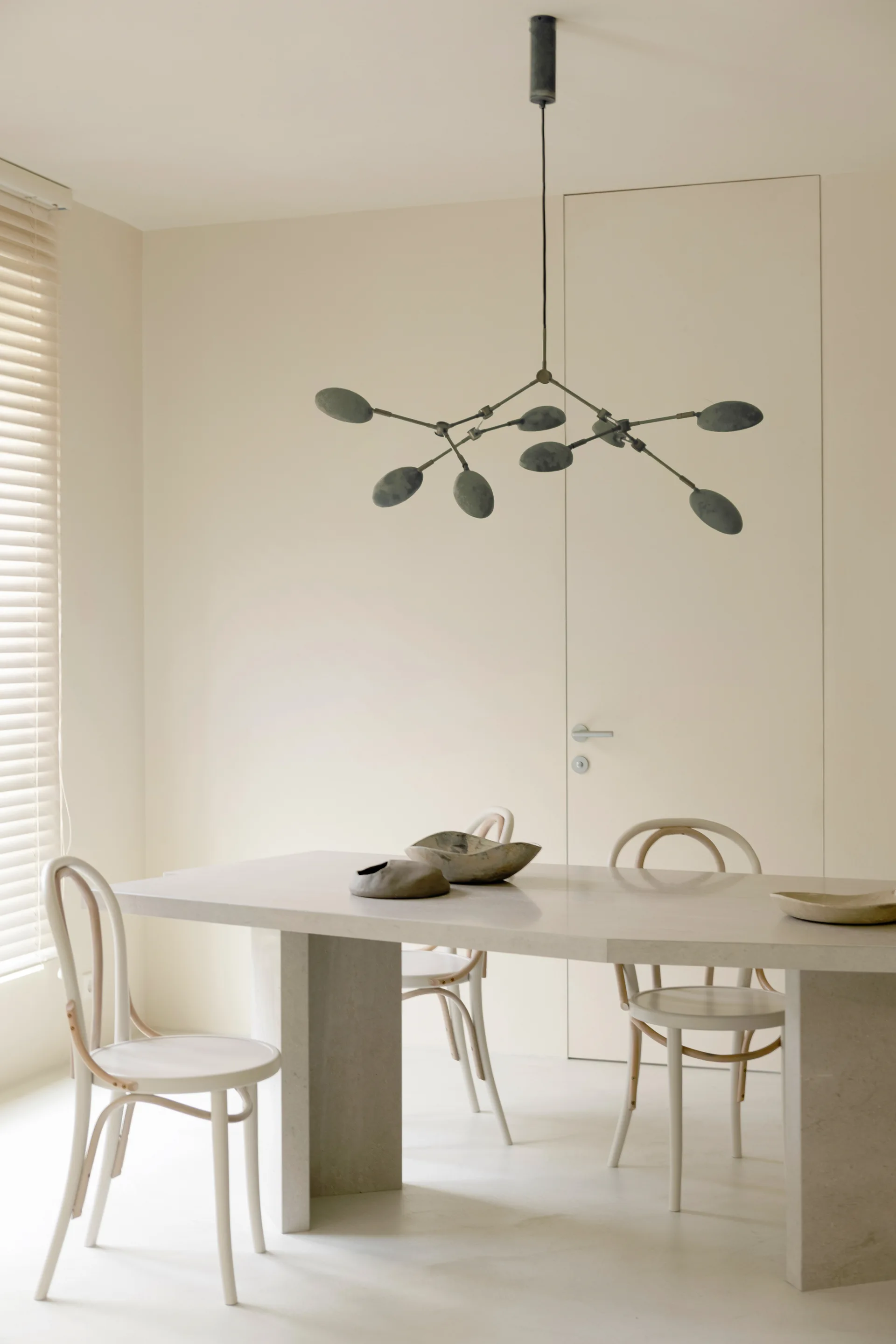 A minimalist dining room features a textured stone table, light wood bentwood chairs, and a dark branched metal pendant light fixture against beige walls.
