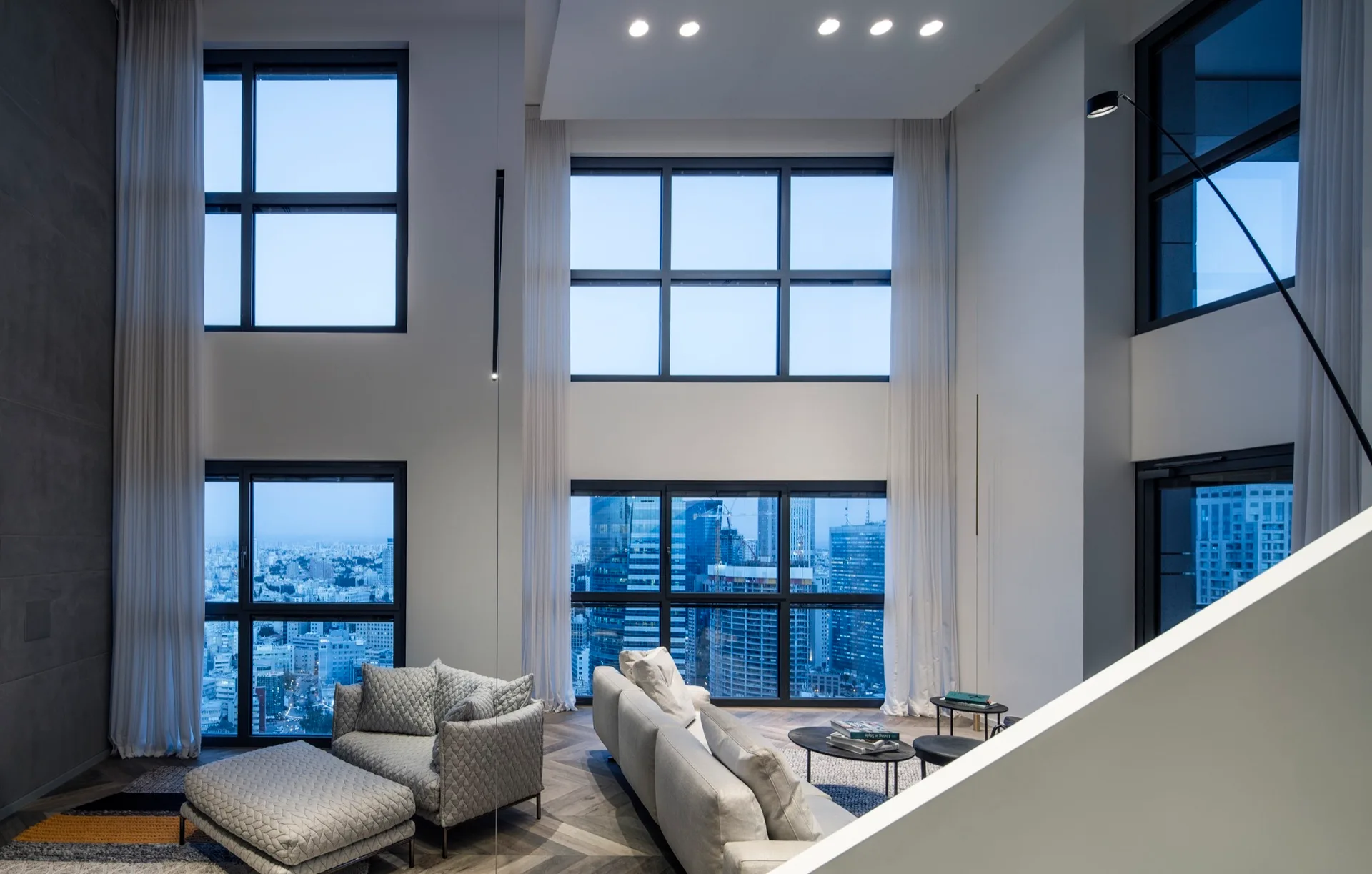 A double-height living room features a grey sectional sofa, a quilted armchair and matching ottoman, chevron wood flooring, and tall stacked windows.