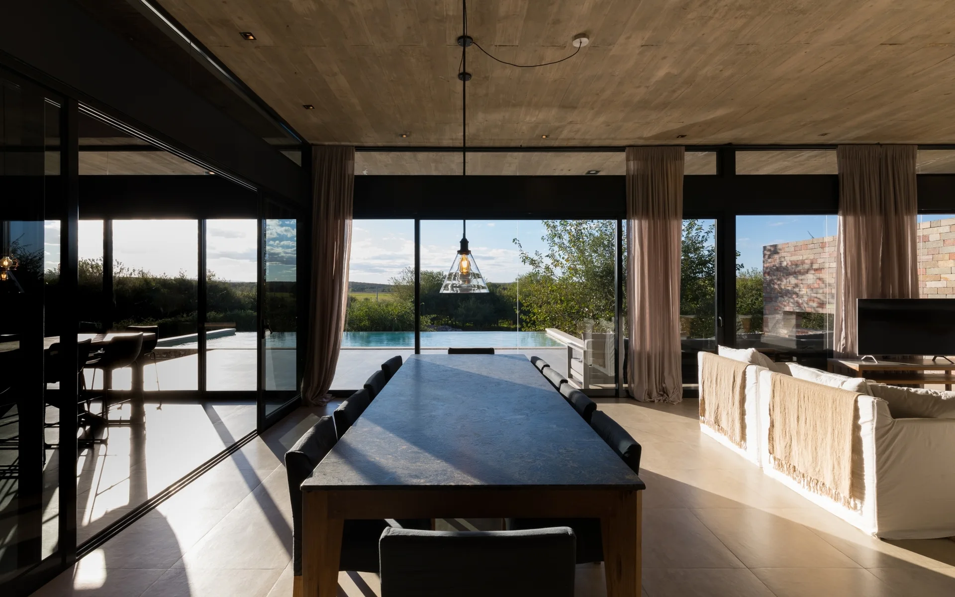 A modern open-plan dining area features a long stone table and dark chairs beneath a raw concrete ceiling, looking out to an outdoor swimming pool.