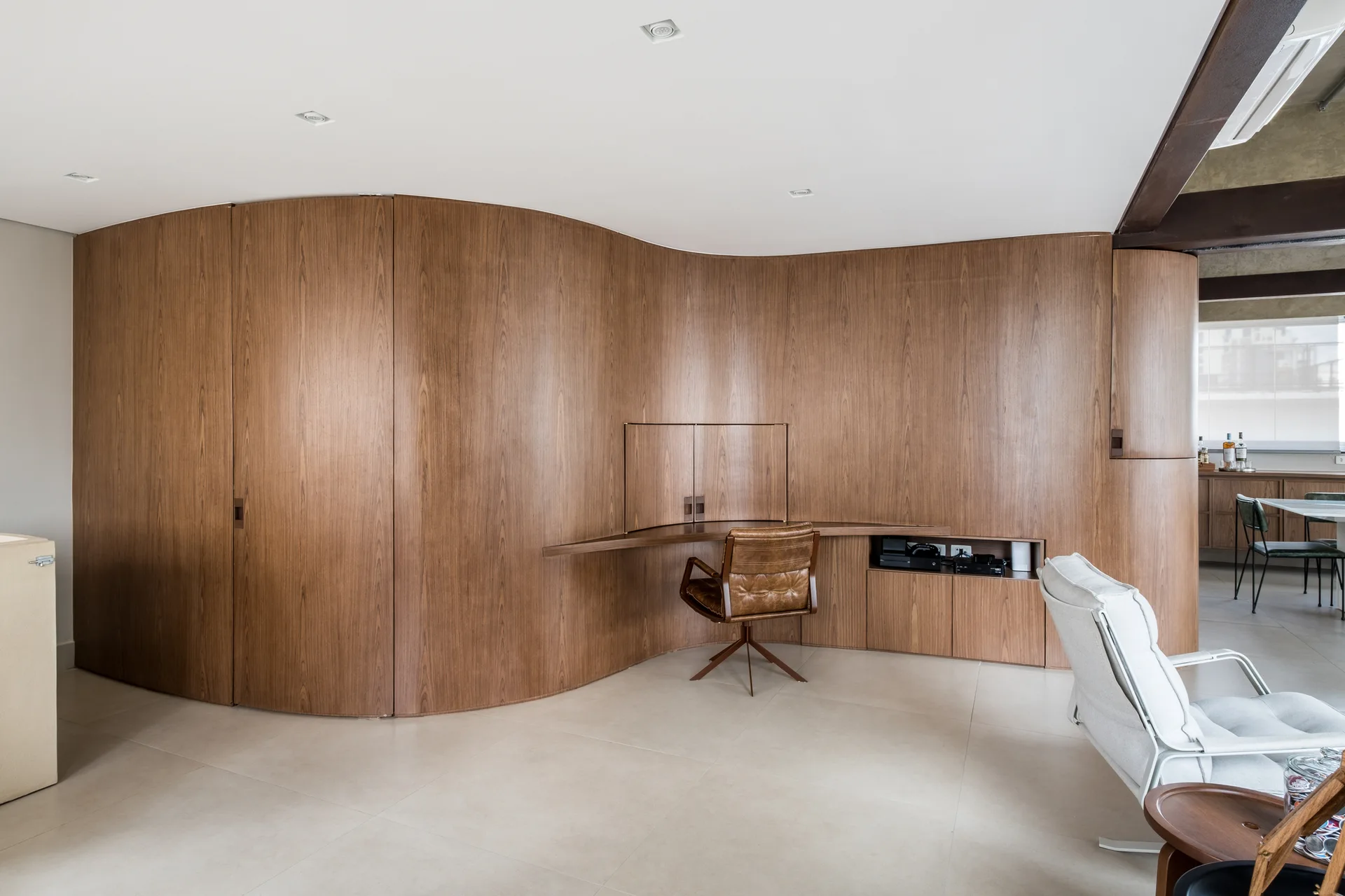 A modern home office features a large curved wood-paneled wall with an integrated desk, a brown leather swivel chair, and beige tiled flooring.