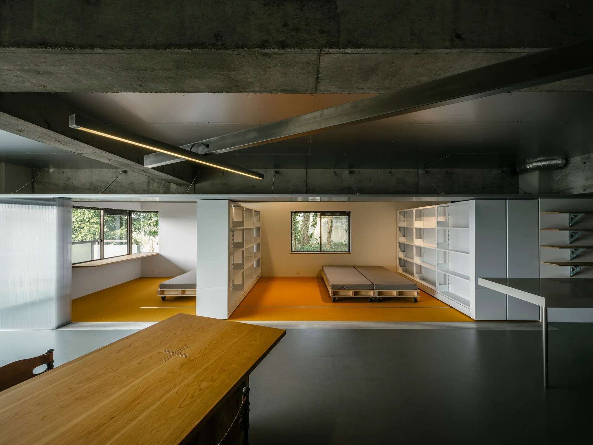 An industrial loft interior with exposed concrete ceilings, a wooden dining table, white modular shelving, and platform beds on vibrant yellow flooring.