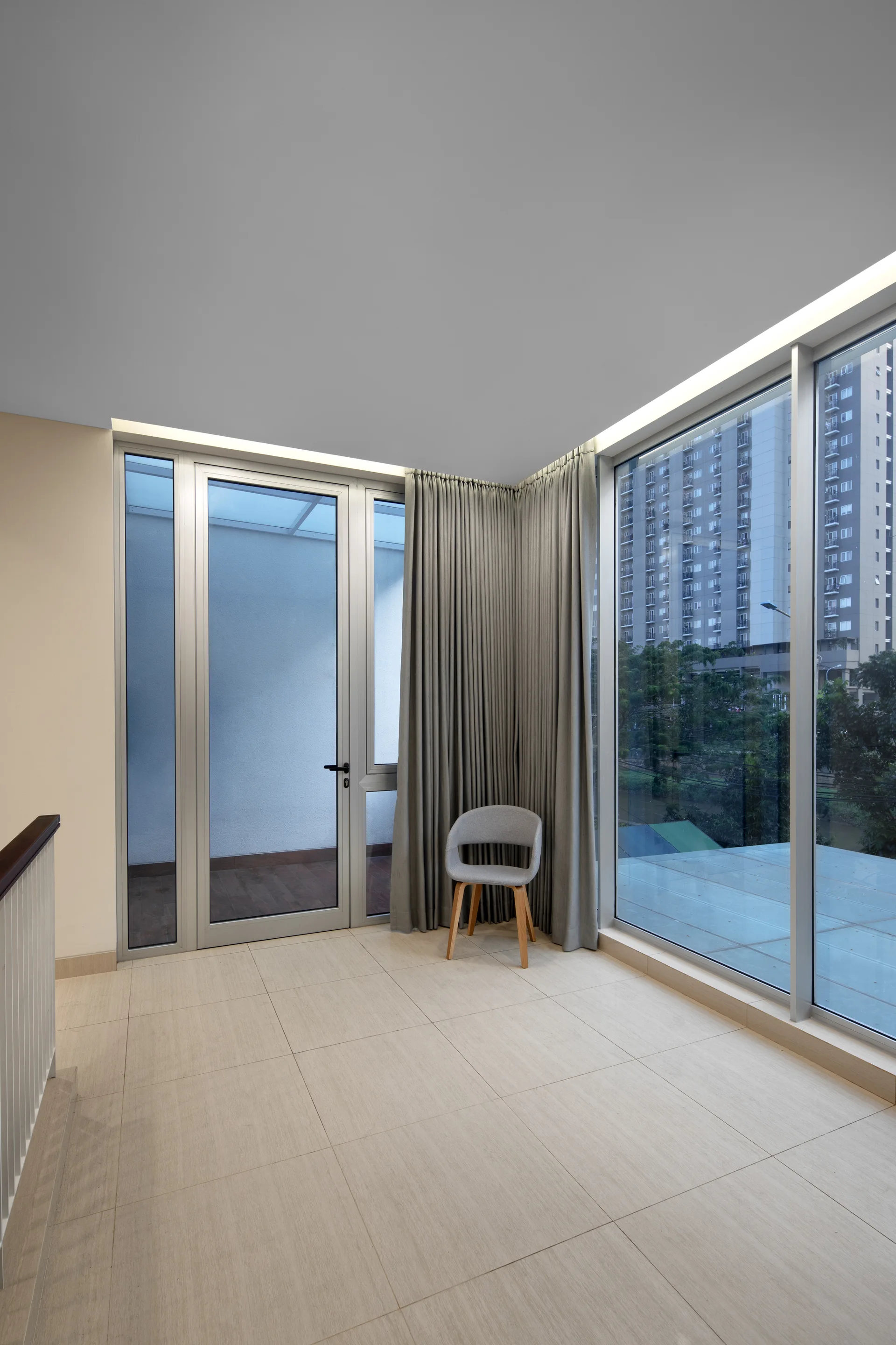 A minimalist staircase landing features light beige floor tiles, large floor-to-ceiling windows with grey drapes, and a single grey accent chair.