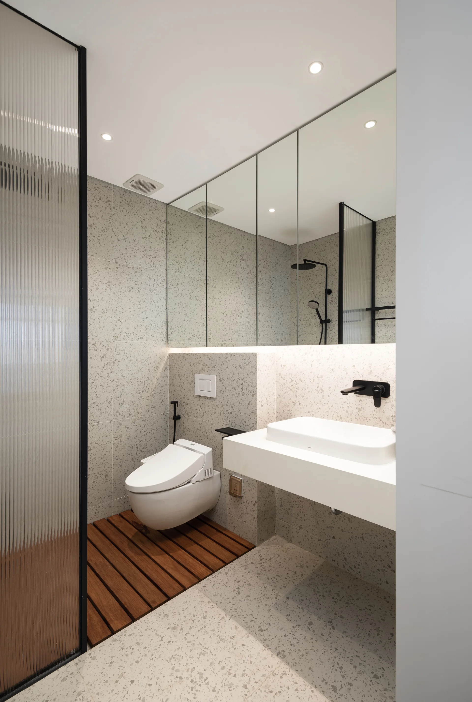 A modern bathroom featuring speckled grey terrazzo walls and floor, a wall-mounted toilet, and a warm wood slat floor inlay.