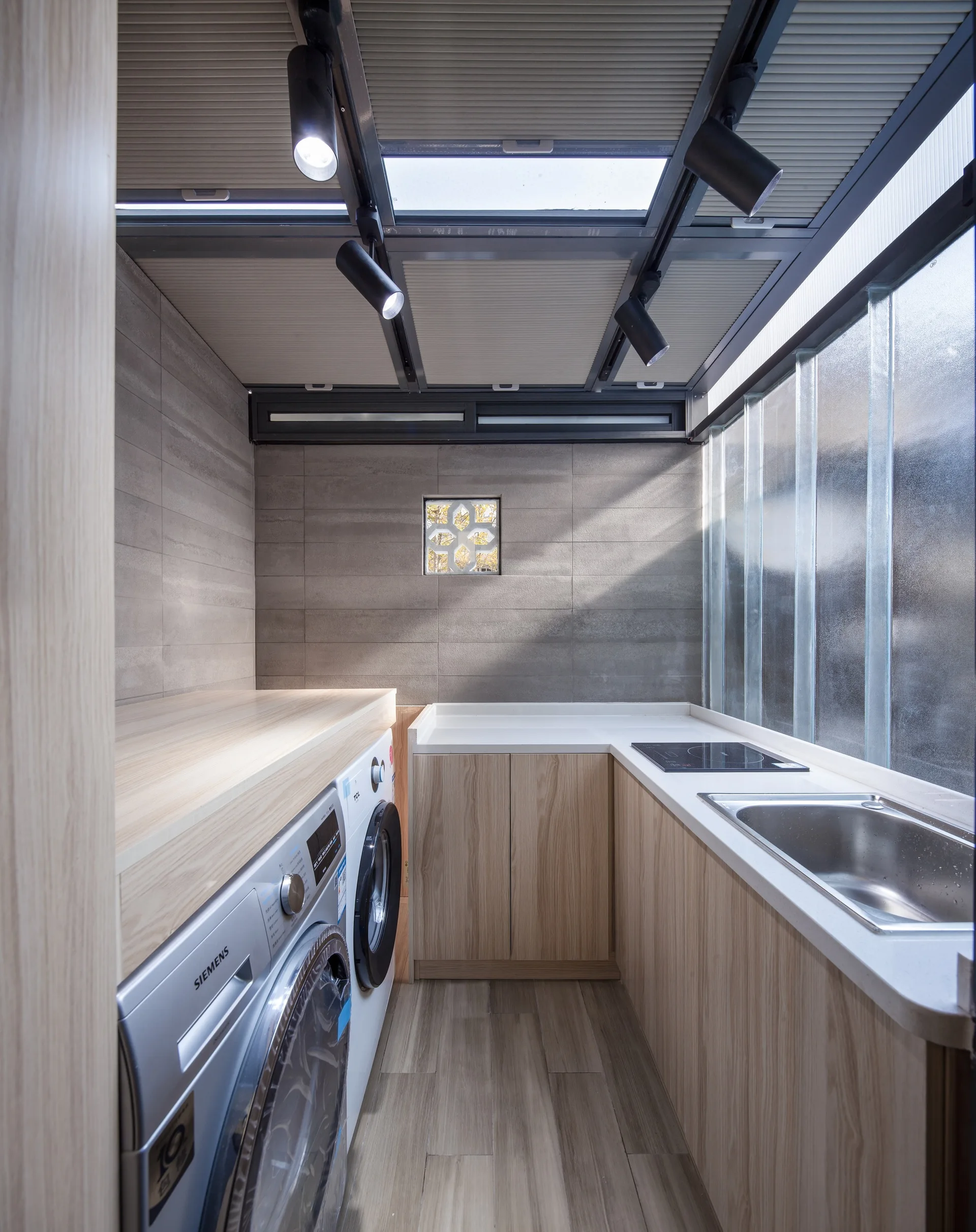 A narrow laundry and utility room with light wood-grained cabinets, a stainless steel sink, a washing machine, and a ceiling skylight.
