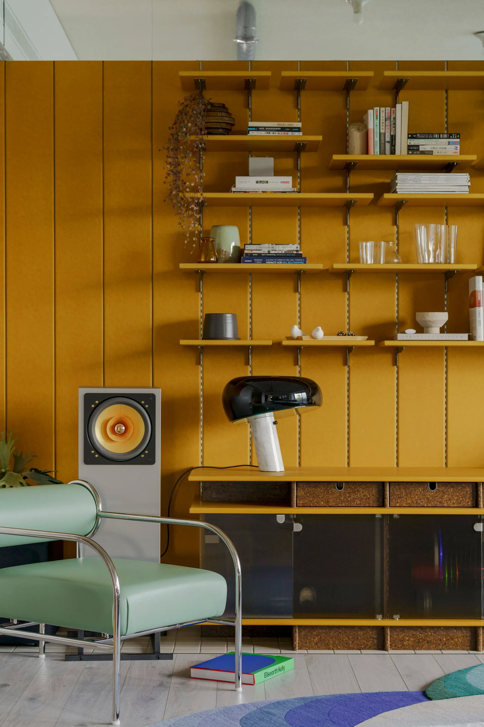 A mint green armchair with a chrome frame sits in front of a mustard yellow paneled wall featuring modular shelving and a low console.