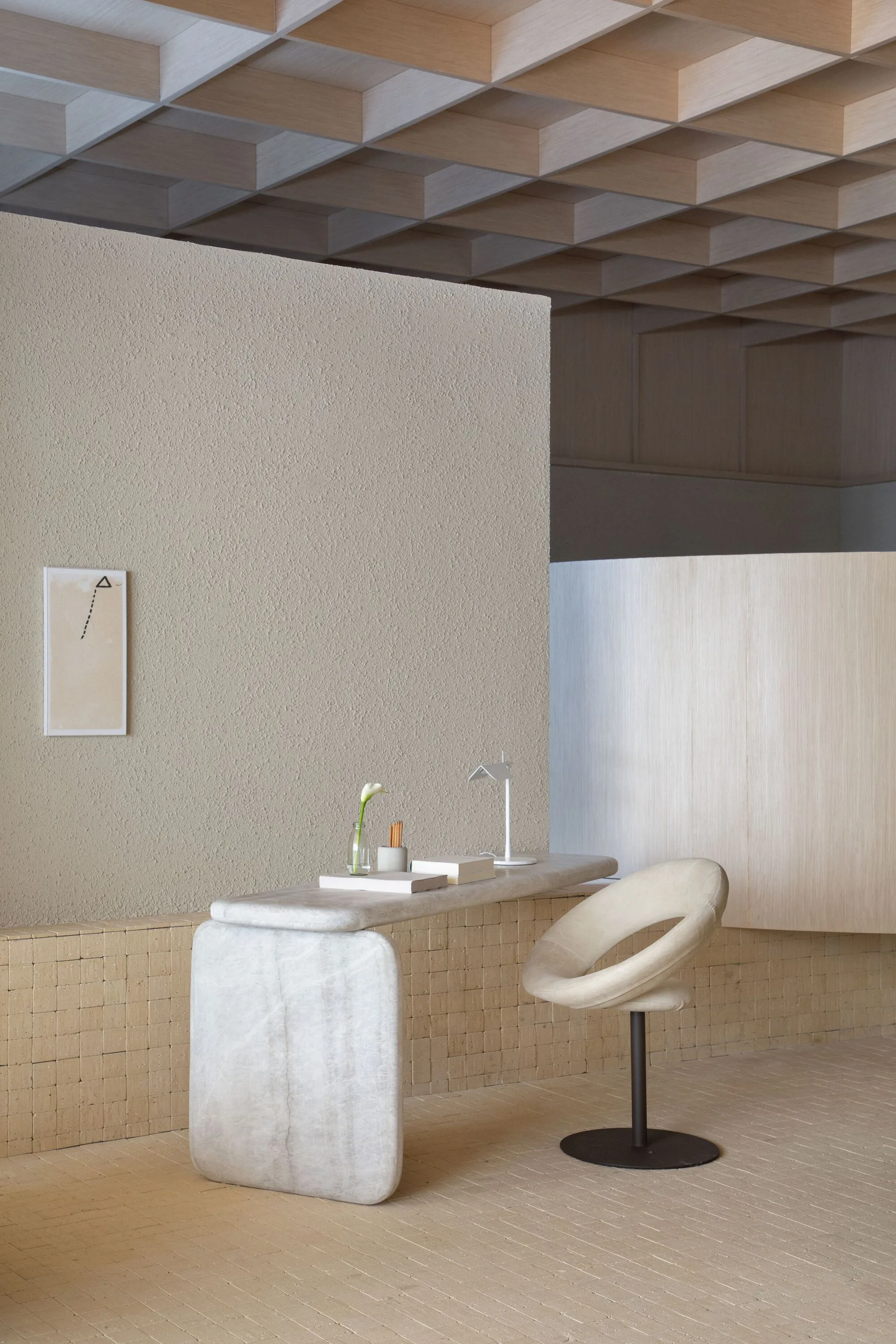 A minimalist home office features a curved stone desk and swivel chair against a textured plaster wall and a wooden coffered ceiling.