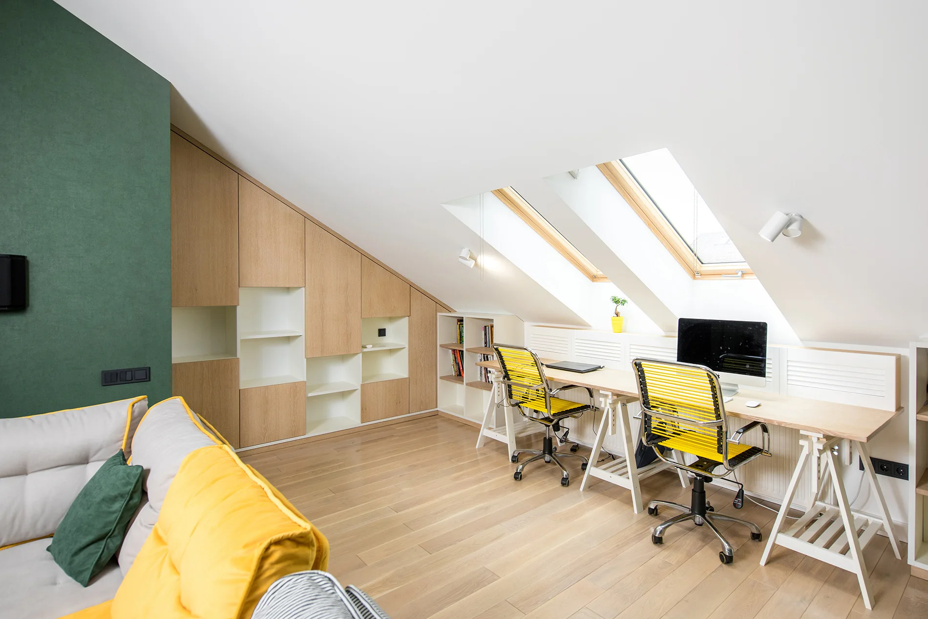 A bright attic home office features a long double desk under dual skylights next to custom wood cabinetry shaped to fit the sloped ceiling.