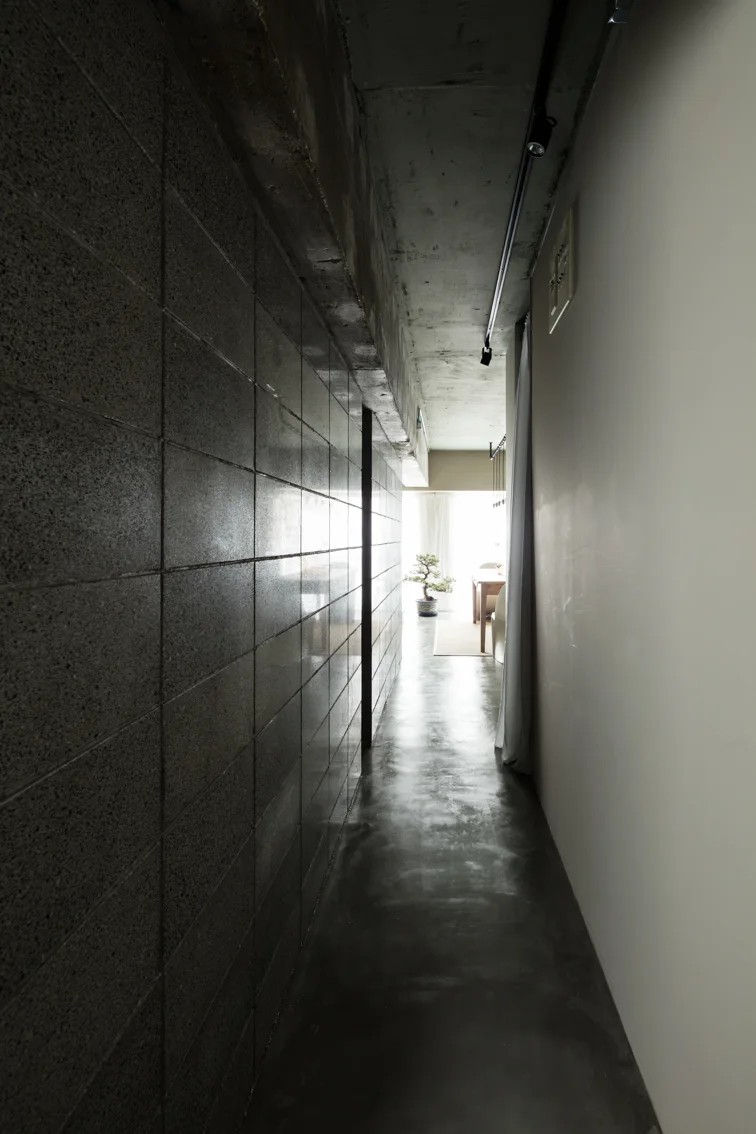 A narrow industrial hallway features a dark concrete block wall, a polished concrete floor, and a bright room with a bonsai tree at the far end.