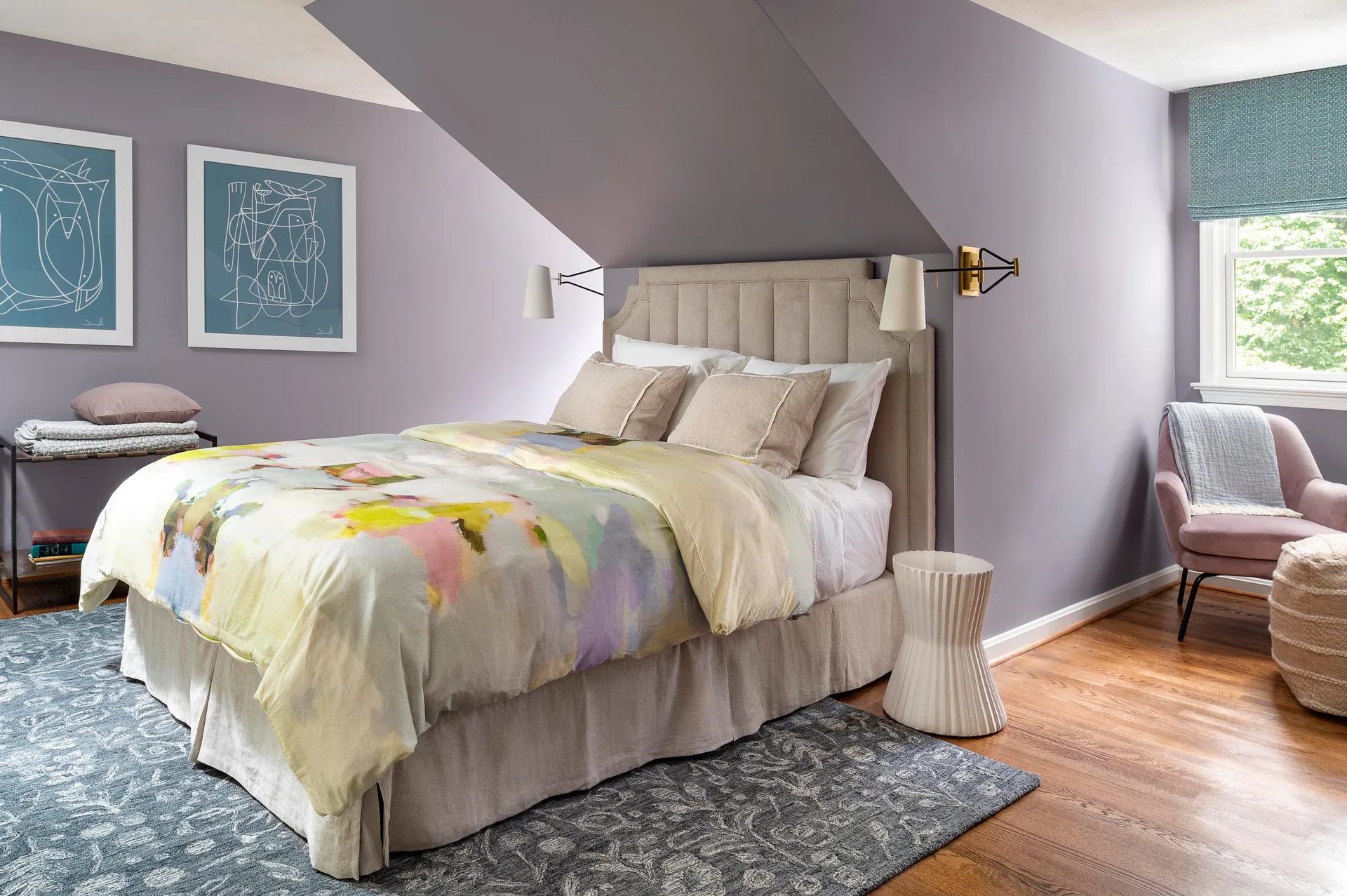 A bedroom with light purple walls and a sloped ceiling features a bed with watercolor bedding and a beige headboard next to a pink accent chair.