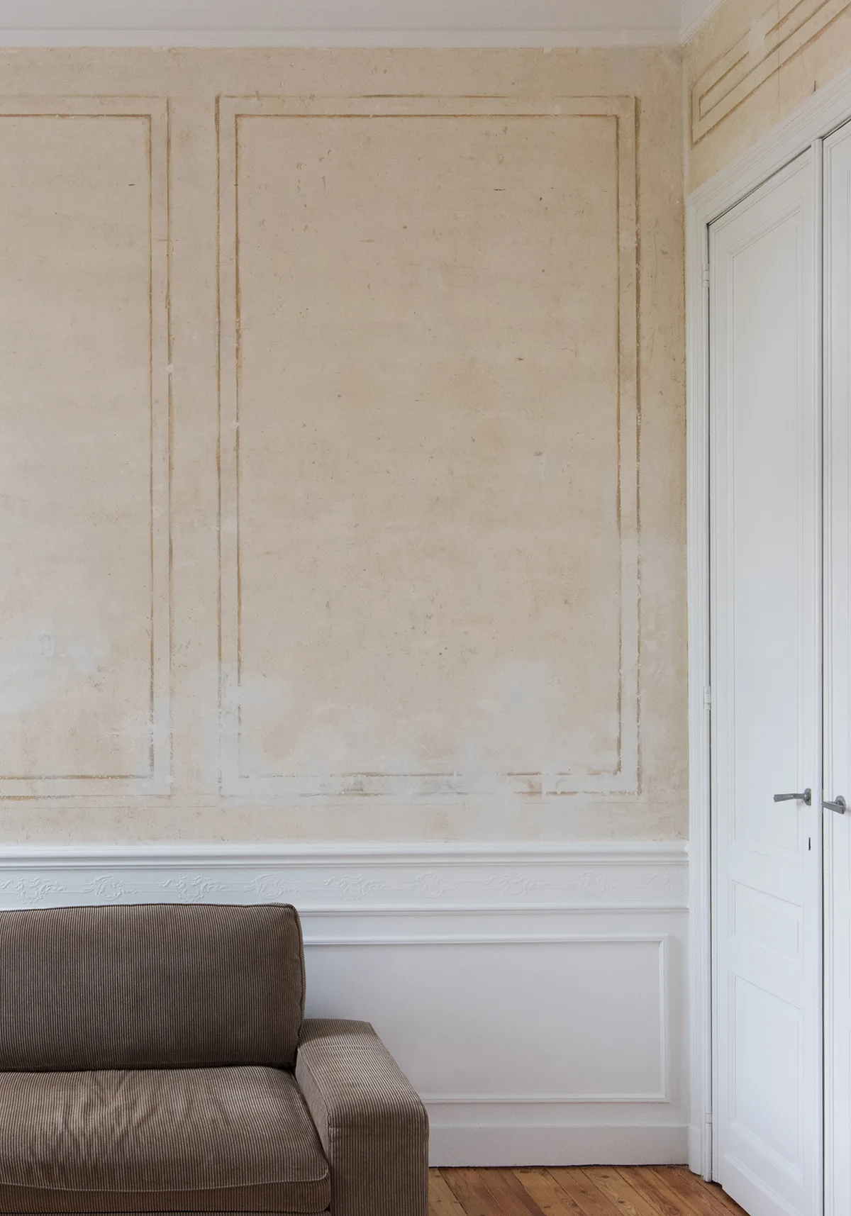 A brown corduroy sofa sits before a beige plaster wall featuring applied moldings, white wainscoting, and a white paneled double door.